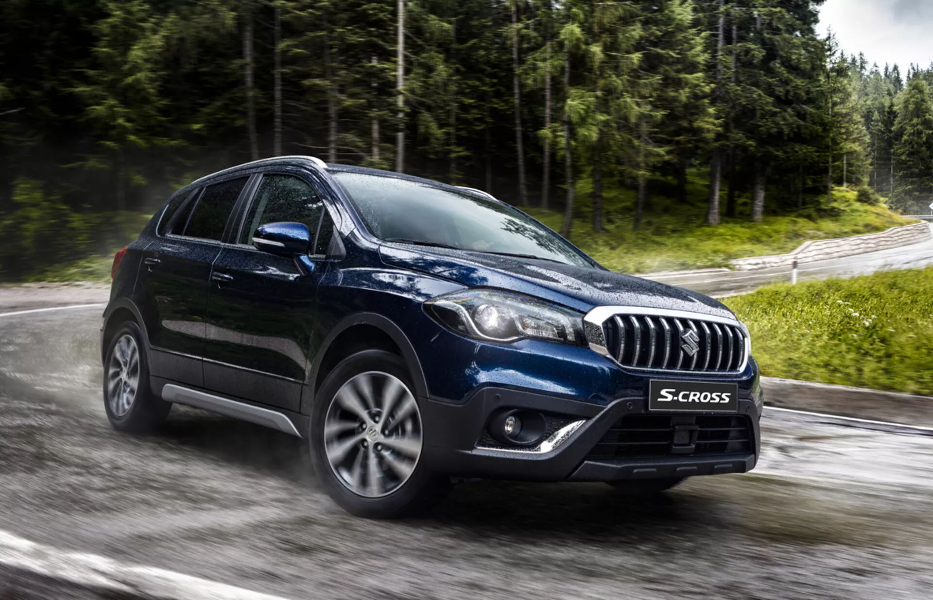 Suzuki S-Cross 2022 zachycený z předního tříčtvrtečního úhlu, zdůrazňující jeho mřížku s chromovými akcenty, moderní LED nebo HID světlomety, slitinová kola s více paprsky a robustní tělo crossoveru na mokré silnici a v zeleni, naznačující pokročilou technologii a schopnost všestranného použití za každého počasí.