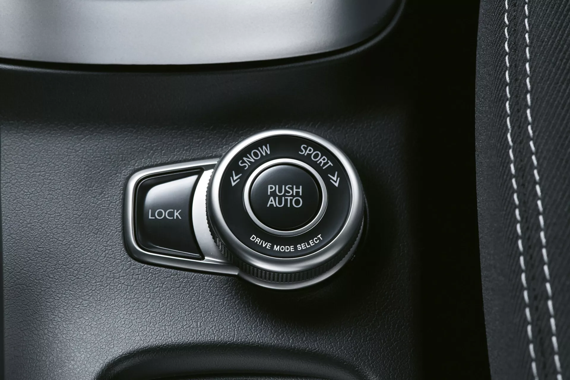 Detailní pohled na interiér Suzuki S-Cross 2022 ukazující volič režimu jízdy označený 'PUSH AUTO' s možnostmi 'SNOW' a 'SPORT' vedle tlačítka 'LOCK', na kvalitním panelu tmavého odstínu, možná s koženou texturou, s viditelným prošíváním.