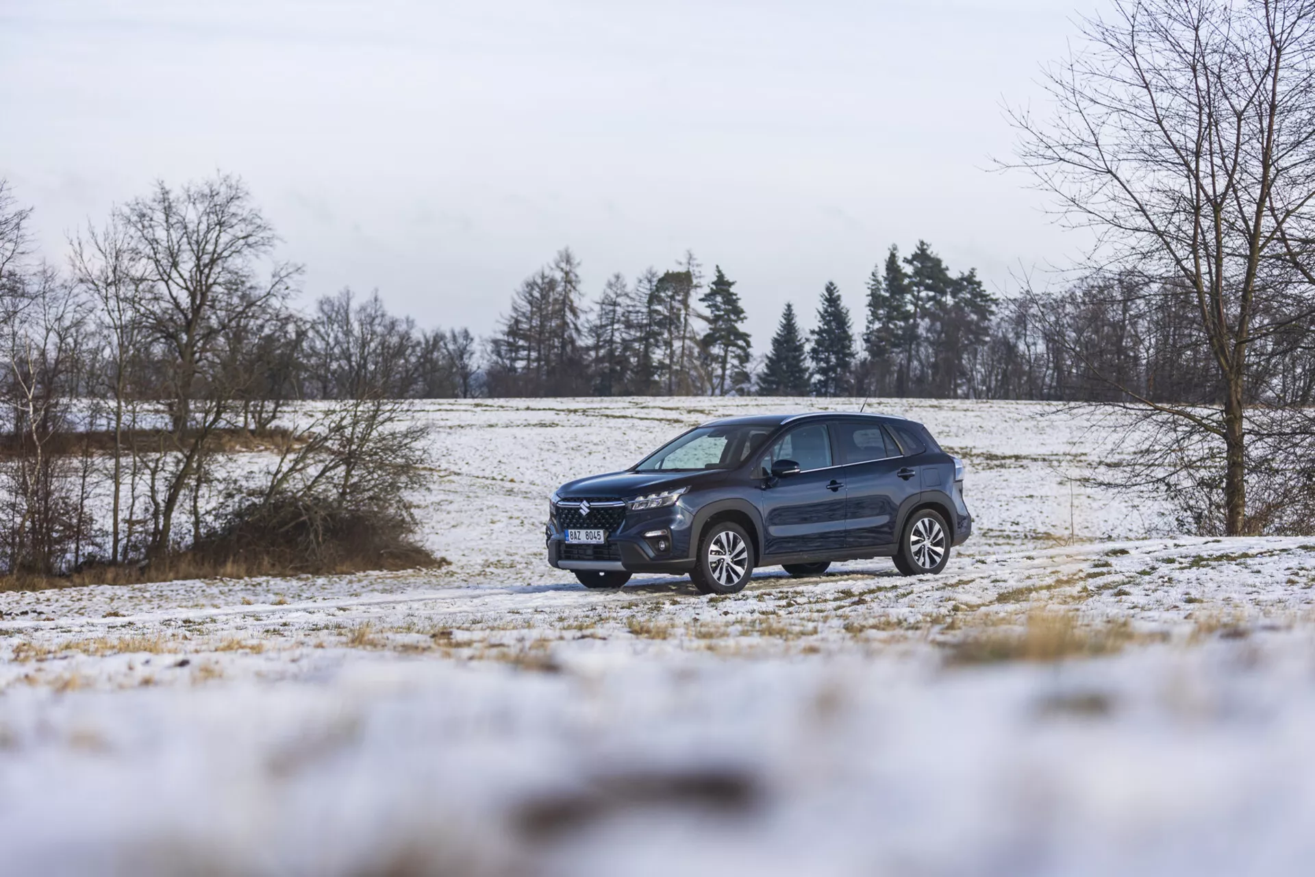 Suzuki S-Cross 2022 v zimní krajině se sněhem, předvádějící svůj dynamický design, pokročilé osvětlení, slitinová kola a střešní nosiče, zdůrazňující jeho stylizaci SUV a schopnost terénní jízdy.