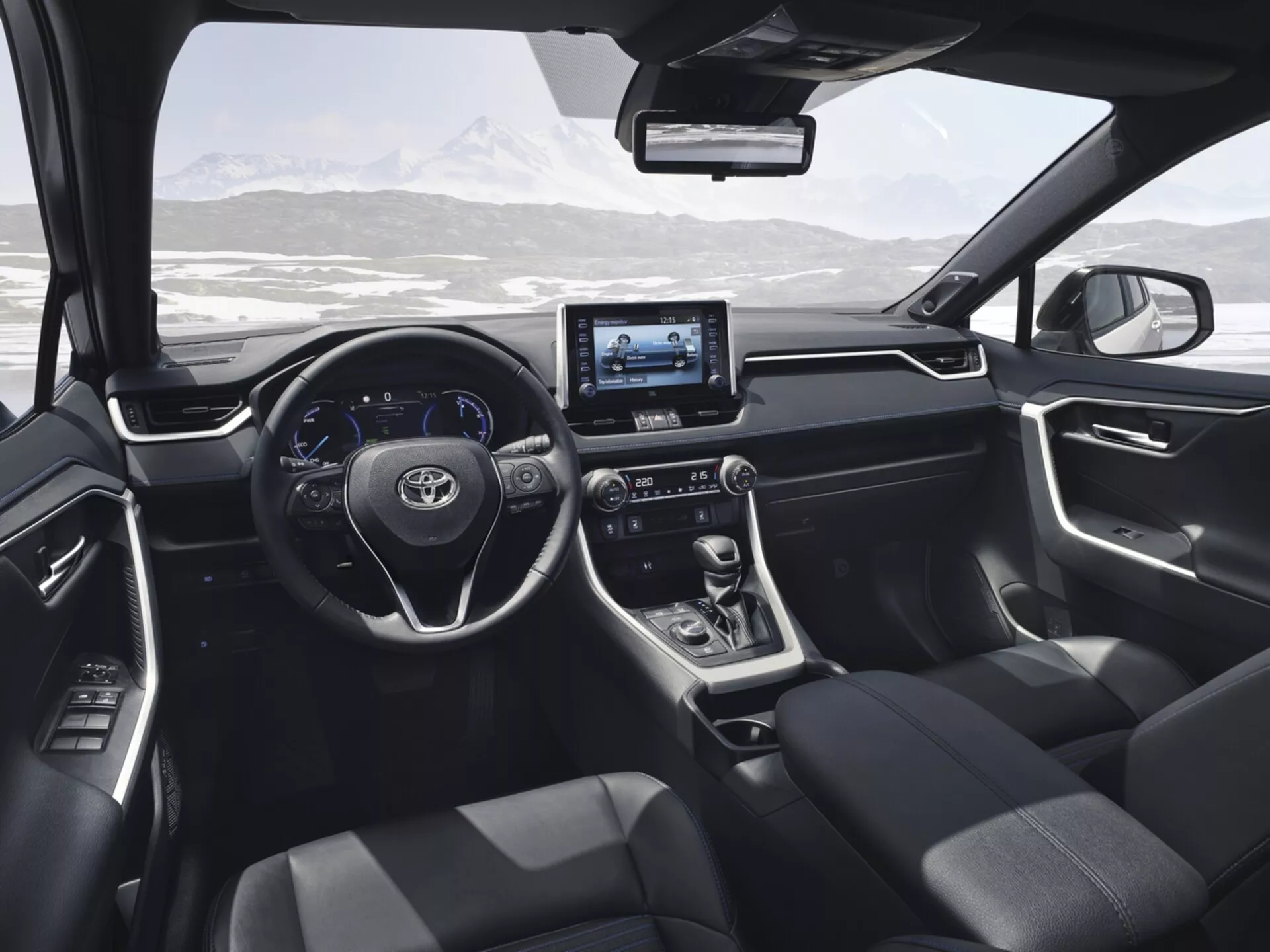 Interiér Toyota RAV4 2019 z pohledu ze zadního sedadla, představující moderní design a technologické prvky včetně volantu s ovládacími tlačítky, digitálního displeje, dotykového infotainment systému, páky automatické převodovky, tmavě čalouněných sedadel a ovládacích panelů na dveřích s výhledem na malebnou zasněženou horskou krajinu skrze čelní sklo.