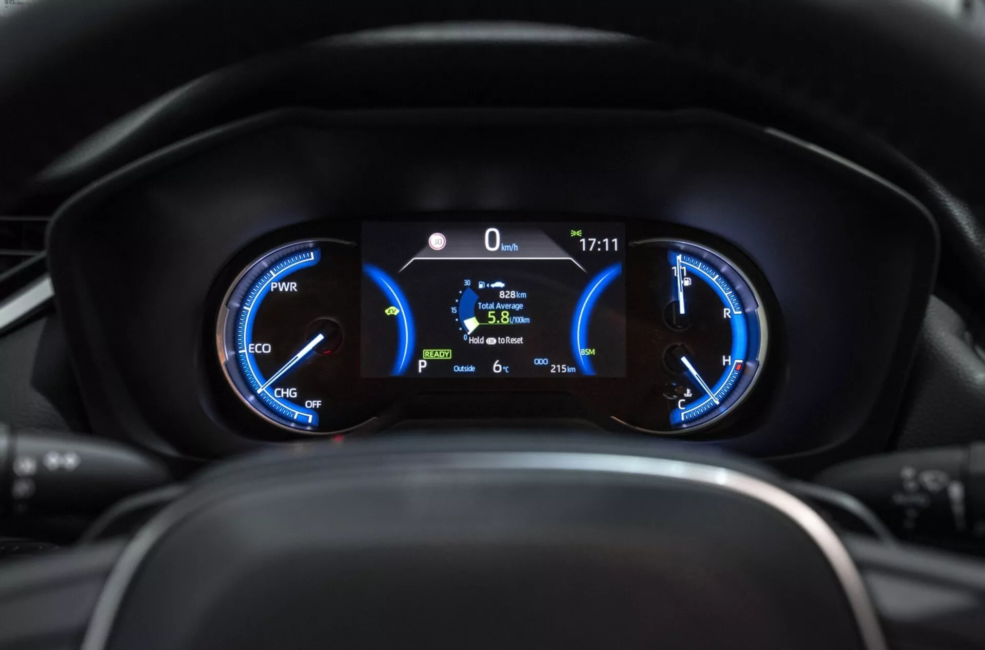 Detailní pohled na přístrojovou desku Toyota RAV4 2019 zobrazující hybridní ukazatele s měřičem výkonu, tachometrem a centrálním digitálním displejem indikujícím stacionární pozici, spotřebu paliva a venkovní teplotu z pohledu řidiče.