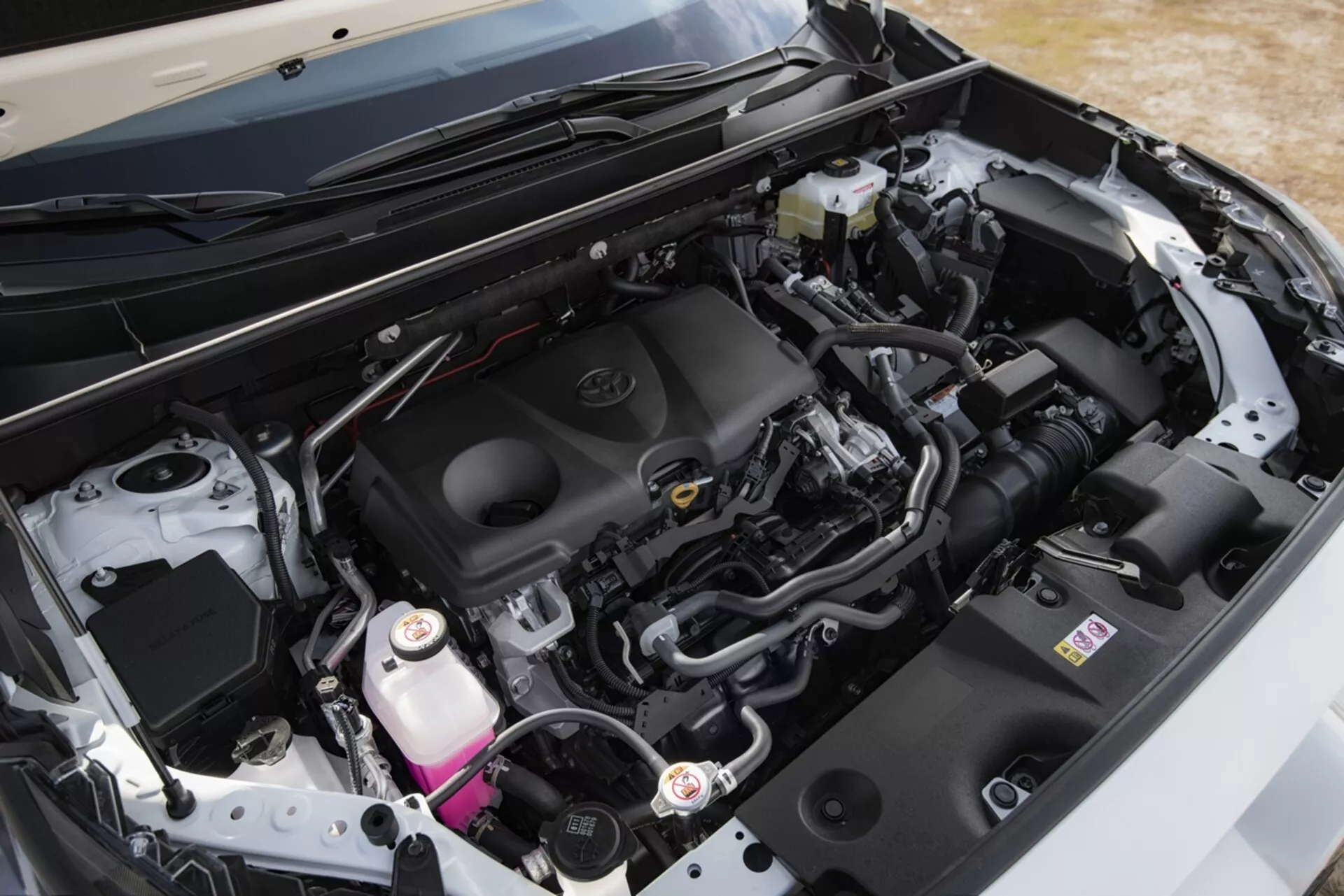 Detailní pohled na motorový prostor Toyota RAV4 z roku 2019, který zdůrazňuje čistý, moderní design a komponenty, jako je kryt motoru s emblémem značky, systém přívodu vzduchu, baterie, nádrž na brzdovou kapalinu a chladicí nádrž, spolu s různými potrubími a svazky kabelů, pod otevřenou kapotou z šikmého úhlu.