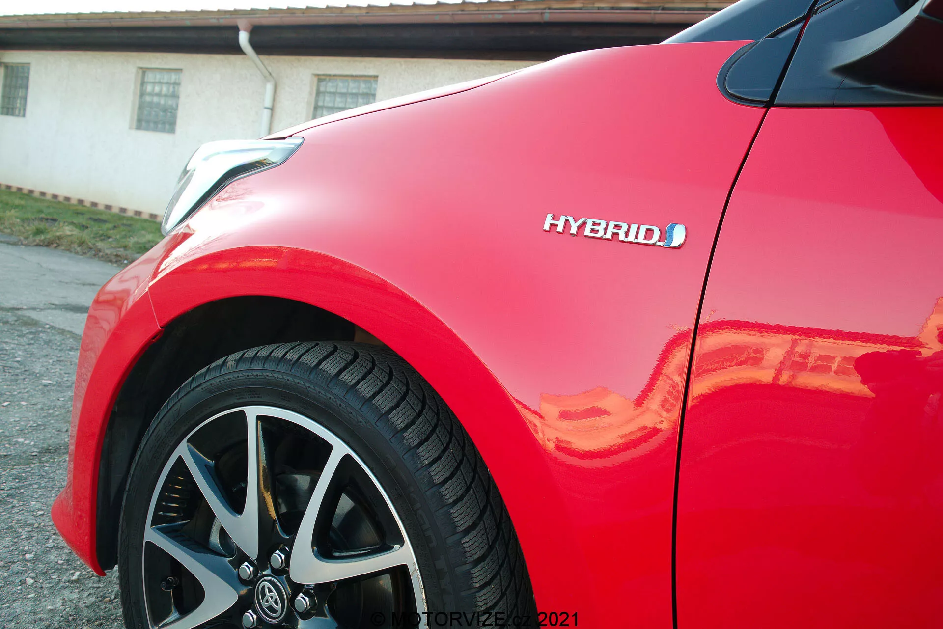 Detailní pohled na červený Toyota Yaris 2020, zaměřující se na přední blatník a vícespeicová slitinová kola. Viditelný je znak hybridu, denukující ekologickou technologii vozidla. Lesklý lak odráží sluneční světlo, zdůrazňující vysokou kvalitu a moderní aerodynamický design vozu.