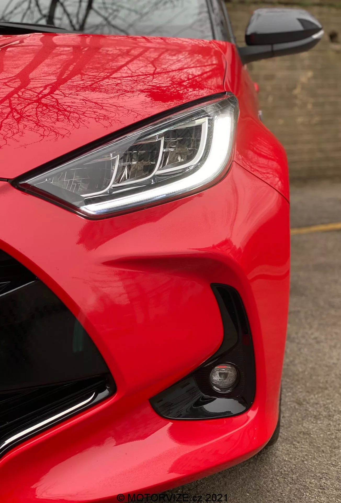 Detailní záběr na Toyota Yaris 2020, ukazující přední část na straně řidiče z nízkého úhlu. Obrázek zdůrazňuje jasně červenou barvu automobilu, elegantní světlomet LED nebo HID s výraznými denními svítilnami a částečně viditelné tělesné zrcátko s integrovaným ukazatelem směru. Aerodynamický design je zdůrazněn vytvarovanými liniemi a křivkami kolem světlometů a kapoty. Kulaté mlhové světlo nebo senzor na předním nárazníku poukazuje na pokročilé bezpečnostní funkce. Absence rozmazání pohybu naznačuje, že automobil je stacionární, a v pravém dolním rohu je viditelný vodoznak 'MOTORZETZ. 2021'.