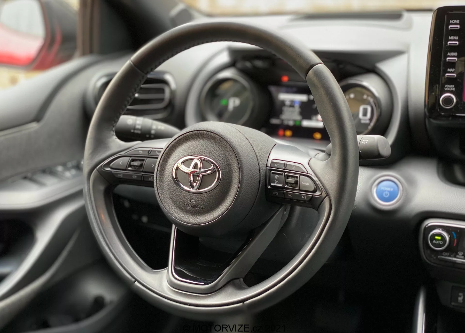 Pohled do interiéru Toyoty Yaris 2020 zaměřený na volant a palubní desku z perspektivy sedadla řidiče. Volant nese logo Toyota a viditelné ovládací prvky pro infotainment a asistenční systémy řidiče. Za ním jsou viditelné části digitálního přístrojového panelu a středové konzoly s ovládacími prvky klimatizace a větracími otvory.