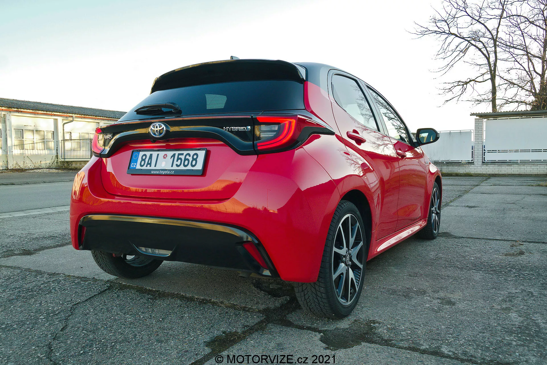 Živě červený osobní automobil Toyota Yaris 2020 fotografovaný zezadu z tříčtvrtinového úhlu, zvýrazňující LED zadní světla, zadní spoiler, znak modelu Hybrid, víceramenné slitinové ráfky a viditelné prvky asistence řidiče, jako jsou parkovací senzory a zadní kamera, zachyceno z trochu pod úrovní výšky auta.
