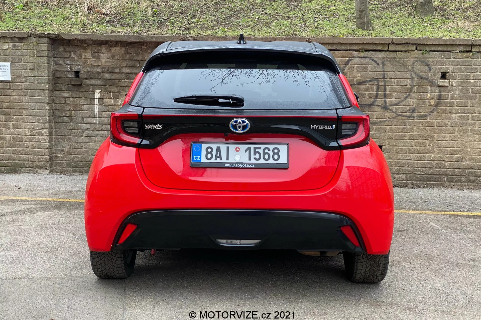 Pohled zezadu na červený Toyota Yaris Hybrid 2020, vycentrovaný a pořízený přímo. Obrázek odhaluje celou šířku vozidla, zobrazující logo Toyota, modelový název 'Yaris' a 'Hybrid', spoiler, střechou namontovanou anténu, brzdové světlo pod spoilerem a výrazná koncová světla. Zadní okno se stěračem a spodní část nárazníku pravděpodobně obsahující senzory nebo odrazky jsou také vidět. Pozadí tvoří cihlová zeď nebo plot a nějaká vegetace, což naznačuje nižší zaparkované auto nebo nižší úhel kamery.