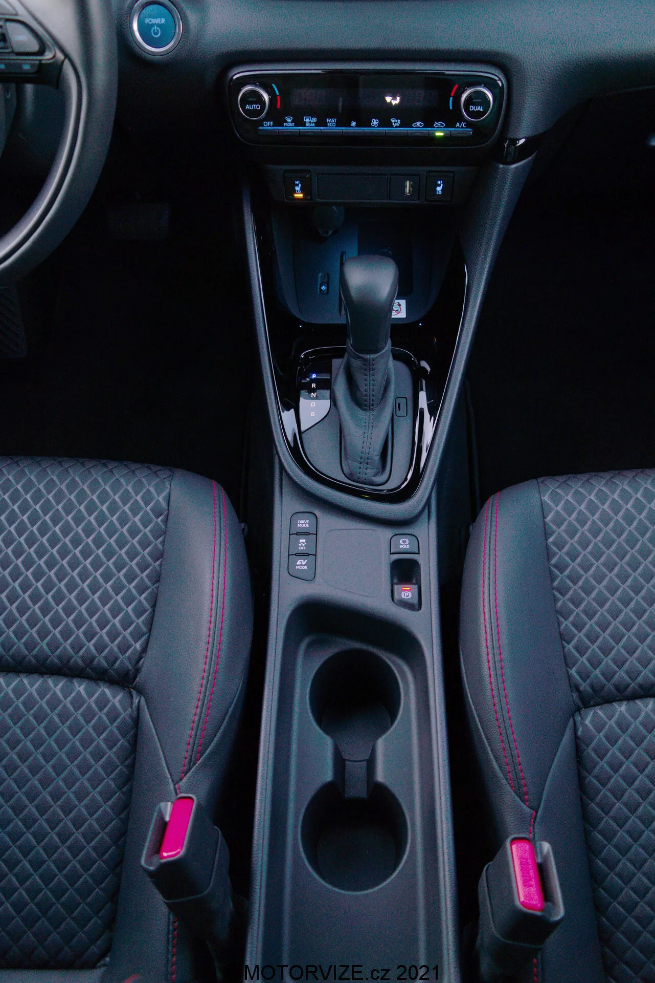 Vertikální záběr interiéru Toyota Yaris 2020 se zaměřením na středovou konzolu, obsahující řadící páku v poloze 'P', elektronické parkovací brzdy, dvě osvětlené držáky nápojů, úložný prostor, digitální klimatizaci, multimediální systém s dotykovou obrazovkou, USB port, ventilační otvory a dvoubarevné přední sedadla s diamantovým prošíváním a červeným lemováním.