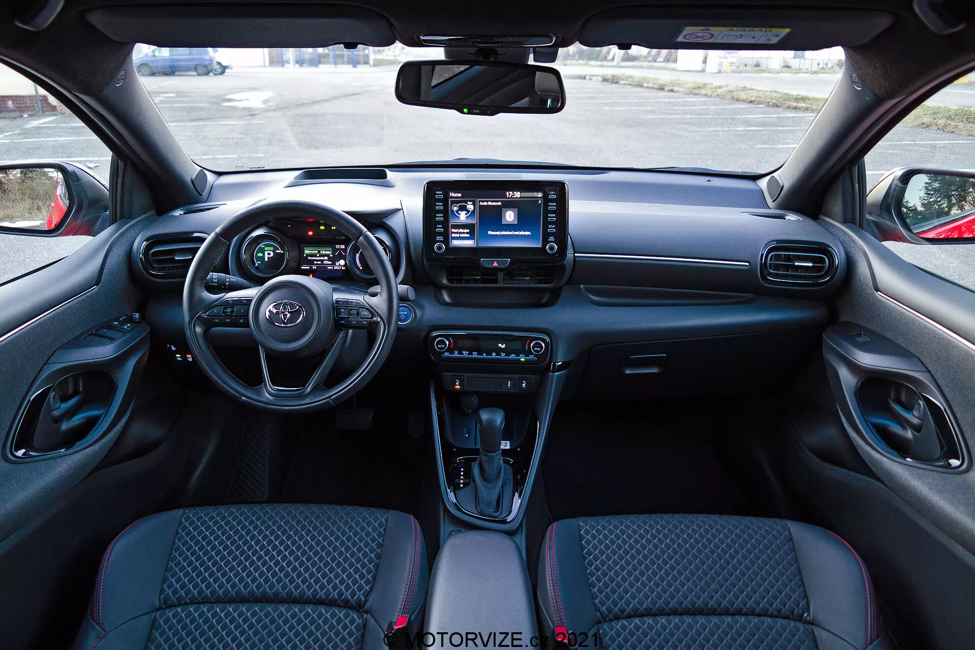 Pohled na interiér vozu Toyota Yaris 2020 z pohledu řidiče, zobrazující moderní palubní desku s volantem, který má integrované ovládací prvky, částečně viditelný přístrojový panel s digitálními obrazovkami a centrální infotainment systém s dotykovým displejem. Palubní deska zahrnuje větrací otvory a knoflíky pro ovládání klimatu, zatímco středová konzola obsahuje ovládací prvky jízdních režimů, automatický řadicí páku s koženým potahem, držáky na nápoje a úložný prostor. Sedadla s látkovým čalouněním mají vzorovaný design a vnitřní části dveří zahrnují ovládací prvky pro elektrická okna a nastavení zrcátek.