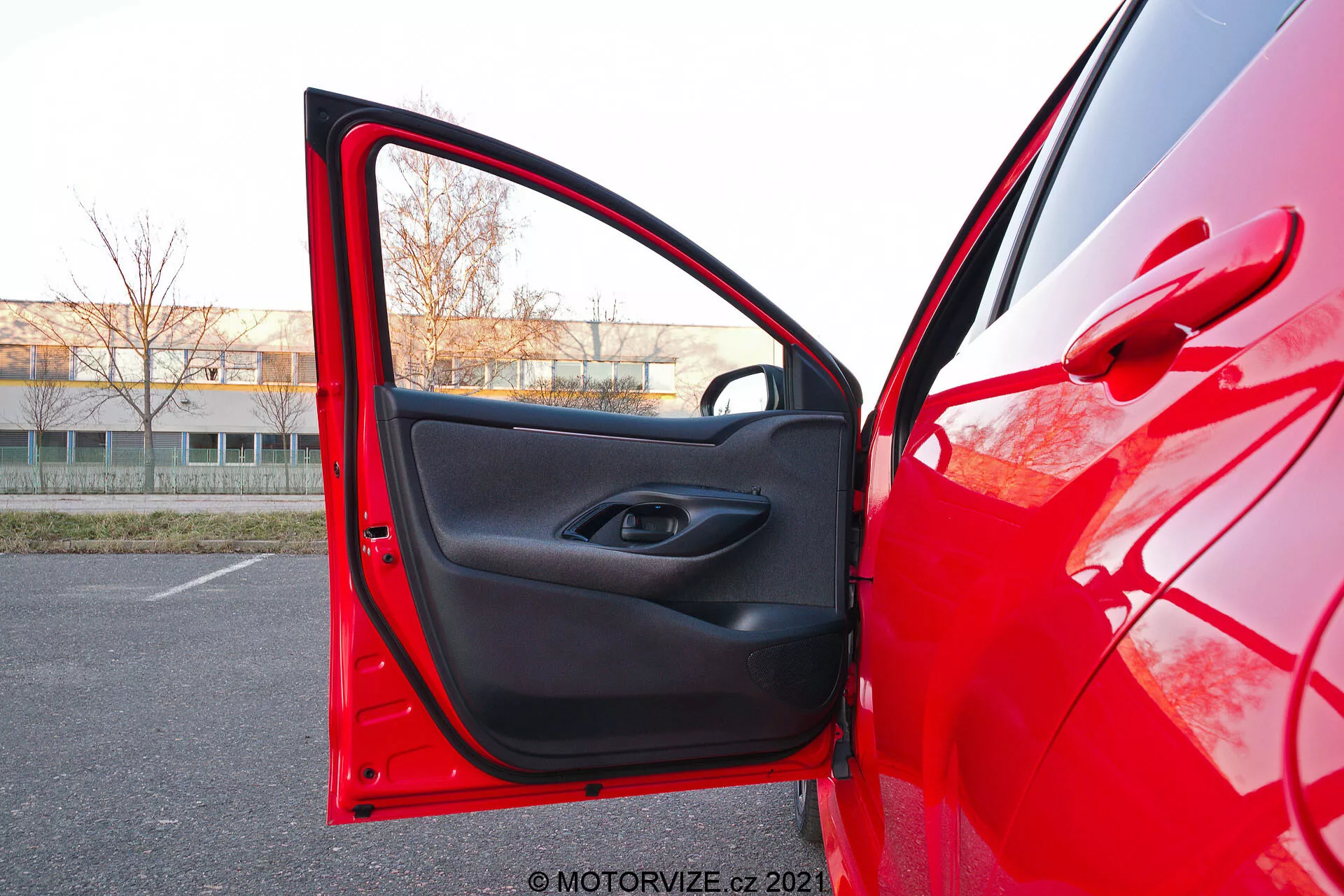 Detailní pohled na červený Toyota Yaris 2020 z boční strany řidiče s otevřenými předními dveřmi, zachycený ze mírně nízkého úhlu. Kamera je umístěna venku, blízko dveří, směřující dovnitř, ukazující jasný červený lak odrážející světlo a černý vnitřní dekor s minimalistickým designem. Dveře mají tlačítko pro ovládání elektrických oken uvnitř rukojeti a pod ní úložnou kapsu.