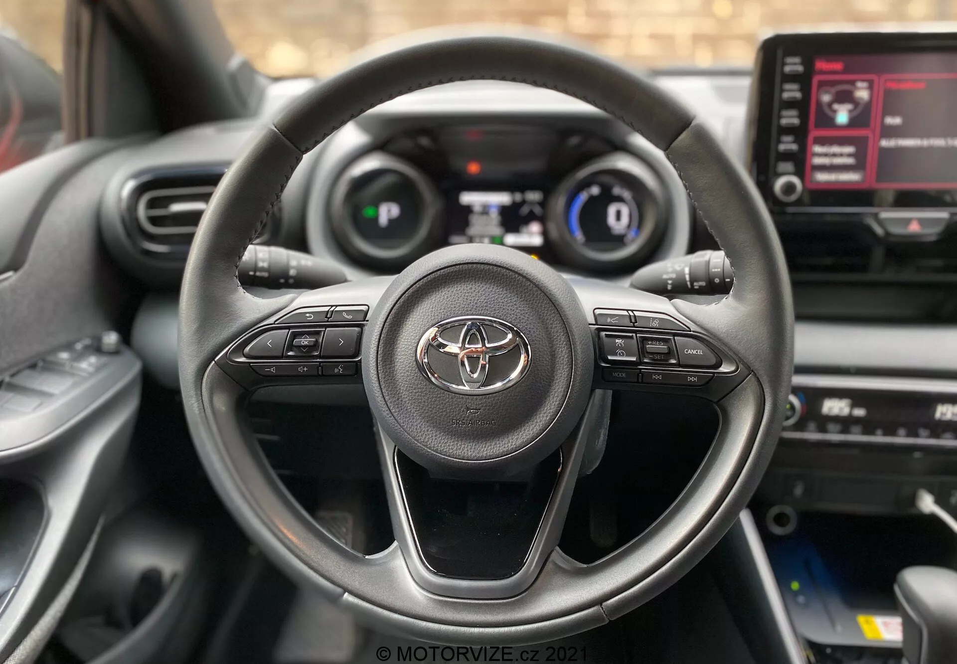 Detailní pohled na interiér Toyoty Yaris 2020 z místa řidiče, zobrazující tříramenný volant s emblémem Toyoty a ovládacími tlačítky pro audio, hands-free volání a tempomat. Digitální přístrojová deska zobrazující rychlost a základní jízdní informace je částečně viditelná. Uprostřed je vidět dotyková obrazovka infotainmentu s nabídkou možností.