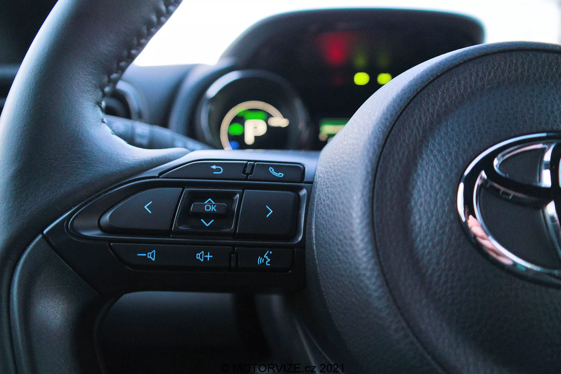 Blízký interiérový pohled na Toyota Yaris 2020 z perspektivy sedadla řidiče, zvýrazňující volant s multifunkčními ovladači a přístrojovým panelem v pozadí. Volant dominuje dolním dvěma třetinám snímku, viditelná tlačítka zahrnují šipky, hlasové ovládání, ovládání hlasitosti a zábavního systému. Částečně viditelný přístrojový panel je osvětlen zeleným digitálním displejem a je rozostřen kvůli nízké hloubce ostrosti. Vnitřek je osvětlen přirozeným světlem, což zdůrazňuje pohodlí a uživatelsky přívětivé rozhraní.