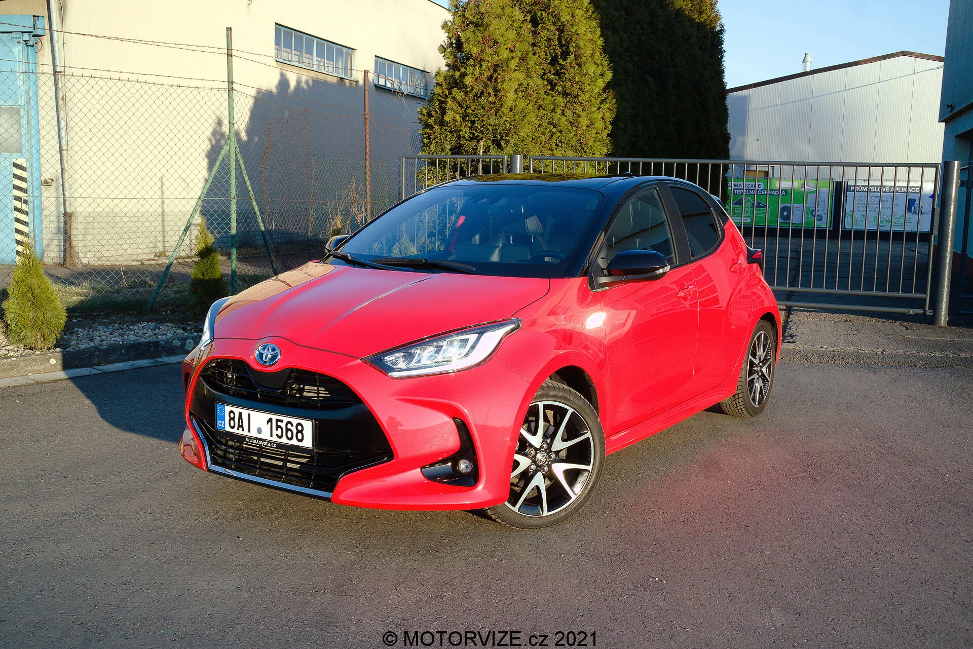 Obrázek jasně červeného hatchbacku Toyota Yaris z roku 2020 zaparkovaného pod úhlem, který ukazuje jak přední, tak boční profil. Design světlometů je ostrý a úhlový, mřížka má voštinový vzor s centrálně umístěným emblémem a přední nárazník má viditelné obrysy s mlhovými světly. Boční profil odhaluje dvoubarevná litá kola, barevně sladěné kliky dveří a zrcátka s ostrými liniemi a obrysy podél boků. Lesklý lak naznačuje nový nebo dobře udržovaný stav. Pozadí zahrnuje jasnou oblohu, průmyslové struktury a měkké stíny, což naznačuje venkovní prostředí, pravděpodobně parkovací prostor poblíž komerčního nebo průmyslového zařízení.