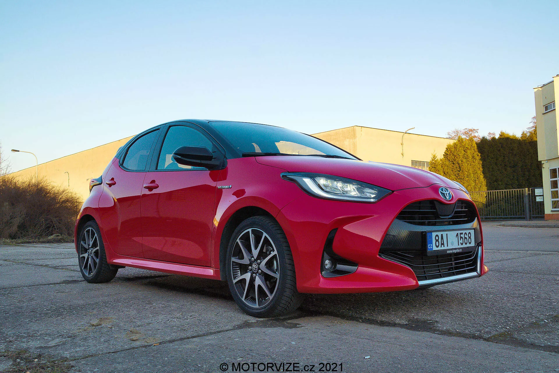 Nízký úhel záběru zepředu tříčtvrtního pohledu na zářivě červený Toyota Yaris 2020 hatchback ukazující ostrá a úhlová LED nebo HID světla, výraznou mřížku s centrálním logem, vícepaprsková hliníková kola, integrované směrovky ve zpětných zrcátkách, čisté reflexní plochy a možná výkonný brzdový systém. Zachyceno ve teplém světle, průmyslovém parkovišti s vodoznakem motorvize.cz 2021.