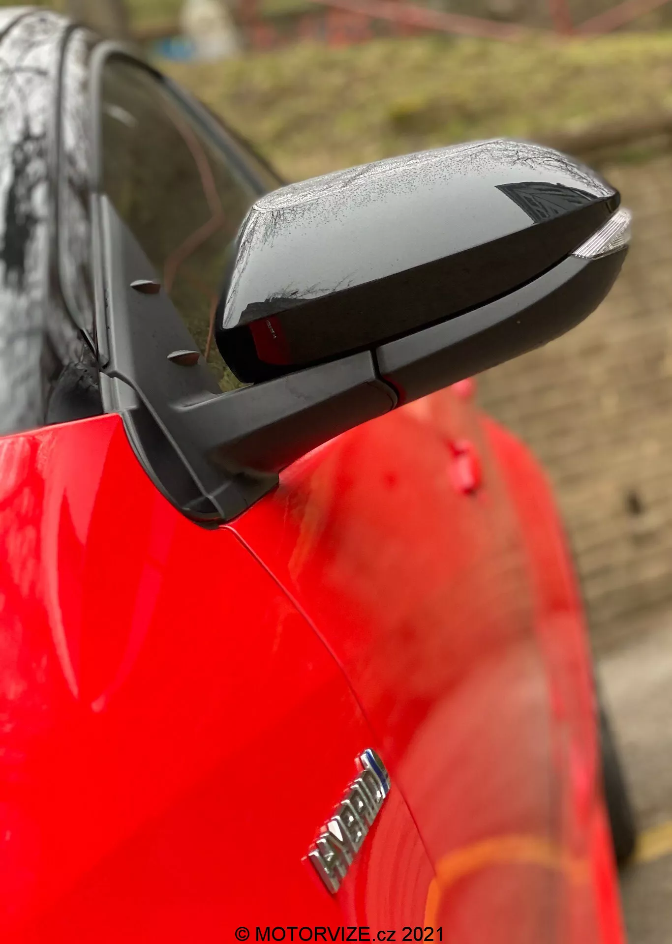 Detailní záběr na vysunuté boční zrcátko na Toyotě Yaris 2020 s elegantním, aerodynamickým černým krytem kontrastujícím s lesklým červeným karoserií. Zrcátko zdůrazňuje technologické prvky jako možné elektrické nastavení, vyhřívání a integrované blinkry, i když konkrétní funkce nejsou potvrzeny. Kapky vody naznačují nedávný déšť nebo mytí auta, zatímco rozmazané pozadí a malá hloubka ostrosti udržují pozornost na zrcátku. Částečně je vidět odznak na předním blatníku, ale ten je mimo ostrost.