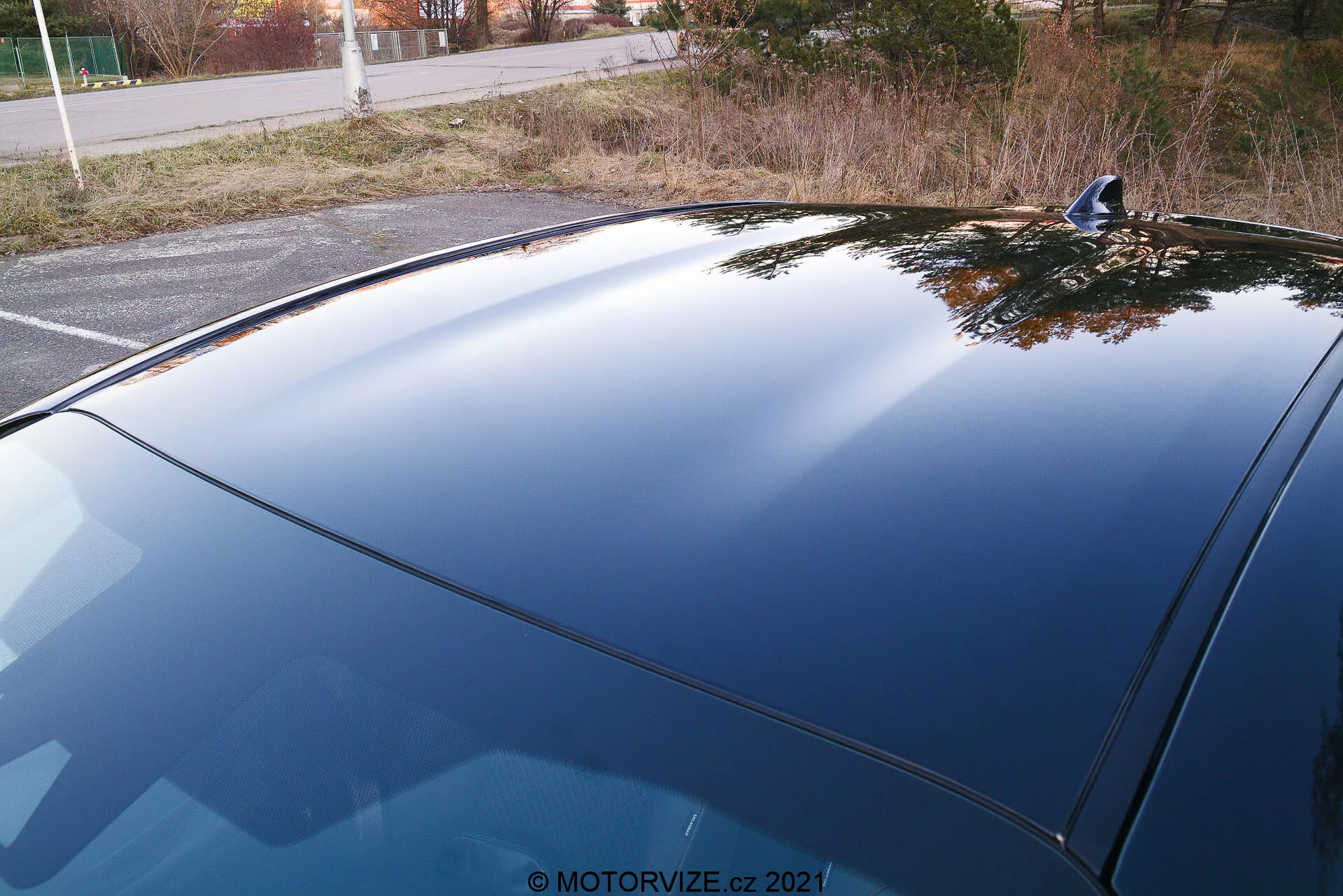 Detailní pohled na zadní část automobilu z nadhledu, zaměřený na zadní okno a část střechy. Auto je zaparkované v tiché krajině s keři a stromy v pozadí. Zadní okno s horizontálními linkami indikuje zabudovaný systém odmrazování. Střecha je pevná a nevypadá na textilní materiál, což naznačuje, že auto není kabriolet. Absence zadního stěrače může znamenat designovou volbu preferující aerodynamiku nebo estetiku. Tónovaná skla poskytují soukromí a ochranu proti UV záření, což je patrné z tmavého zadního okna. Celkový stav automobilu působí dobře udržovaným a čistým dojmem, s lesklým lakem a bez viditelných škrábanců nebo poškození.