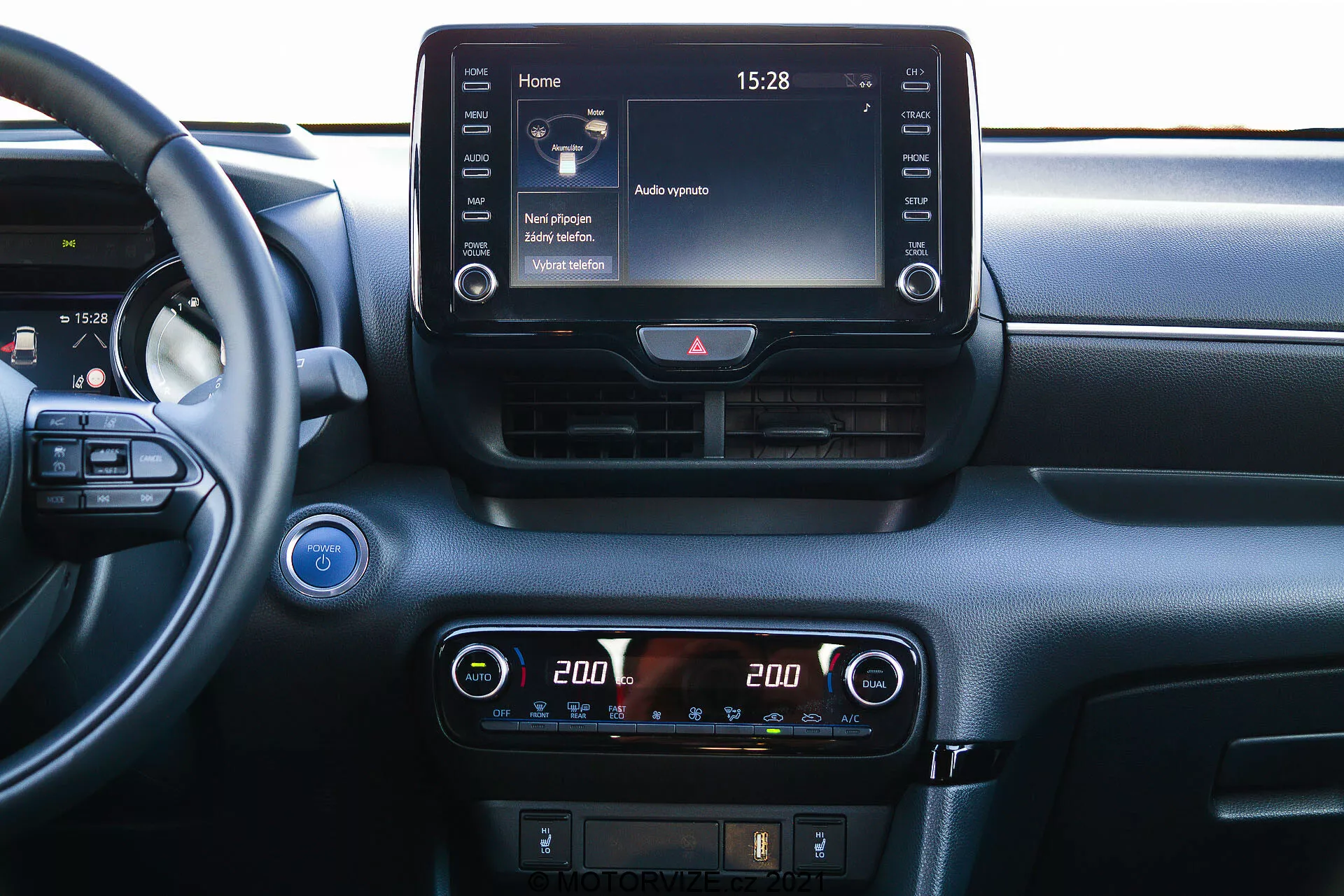 Blízký pohled na centrální palubní desku Toyoty Yaris 2020 z interiéru. Částečně viditelný volant vlevo naznačuje úhel pohledu z místa spolujezdce nebo ze středu. Palubní deska obsahuje výrazný infotainment dotykový displej na vrcholu centrální konzole, zobrazující domovskou obrazovku s různými ikonami a čas '15:28'. Pod ním je digitální displej klimatizace s nastavením dvouzónové teploty, každá strana nastavena na '20.0' stupňů Celsia. Pod klimatizací jsou vidět další ovládací prvky pro odmlžování a možná řízení asistence řidiče. Nad infotainment displejem se nachází dvě horizontální větrací otvory s tlačítkem varovných světel mezi nimi. Na levé straně je částečně viditelné tlačítko start/stop, což naznačuje technologii bezklíčového zapalování. Vlevo nahoře se nachází mřížka reproduktoru. Materiál palubní desky obsahuje hladké i texturované prvky, což naznačuje zaměření na pohodlí a technologii.