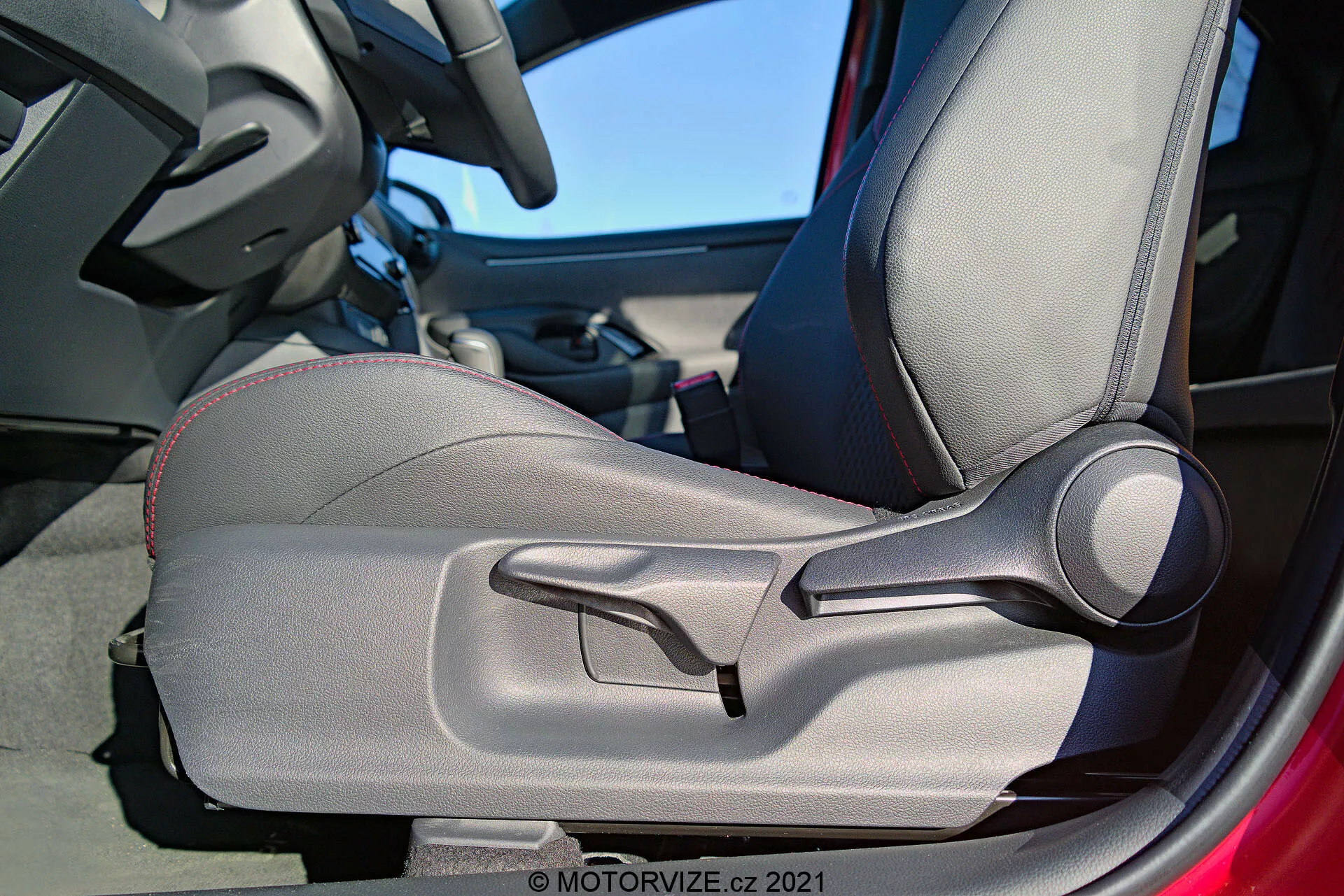 Vnitřní detailní záběr na Toyota Yaris 2020 zaměřený na dveře a sedadlo řidiče, zvýrazňující dvoubarevné čalounění, dekorativní šití, tvarovaný design pro ergonomickou podporu, ručně nastavitelné boční opěrky a výraznou páku pro ruční nastavení sedadla. Dveřní rám je částečně viditelný a naznačuje, že dveře jsou otevřené.