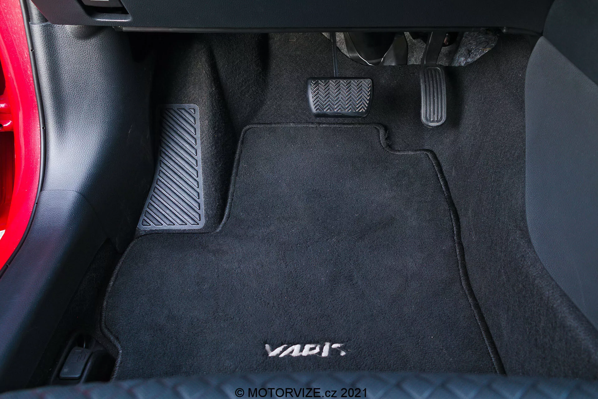 Detailní pohled na prostor pro nohy na straně řidiče ve vozidle, zdůrazňující tři pedály s kovovým povrchem a gumovými vložkami pro manuální převodovku na černé kobercové podložce s logem 'VAG'. Levý pedál je spojka, střední je brzda a pravý je plynový pedál. Částečně viditelná je spodní část palubní desky a část dveří auta.