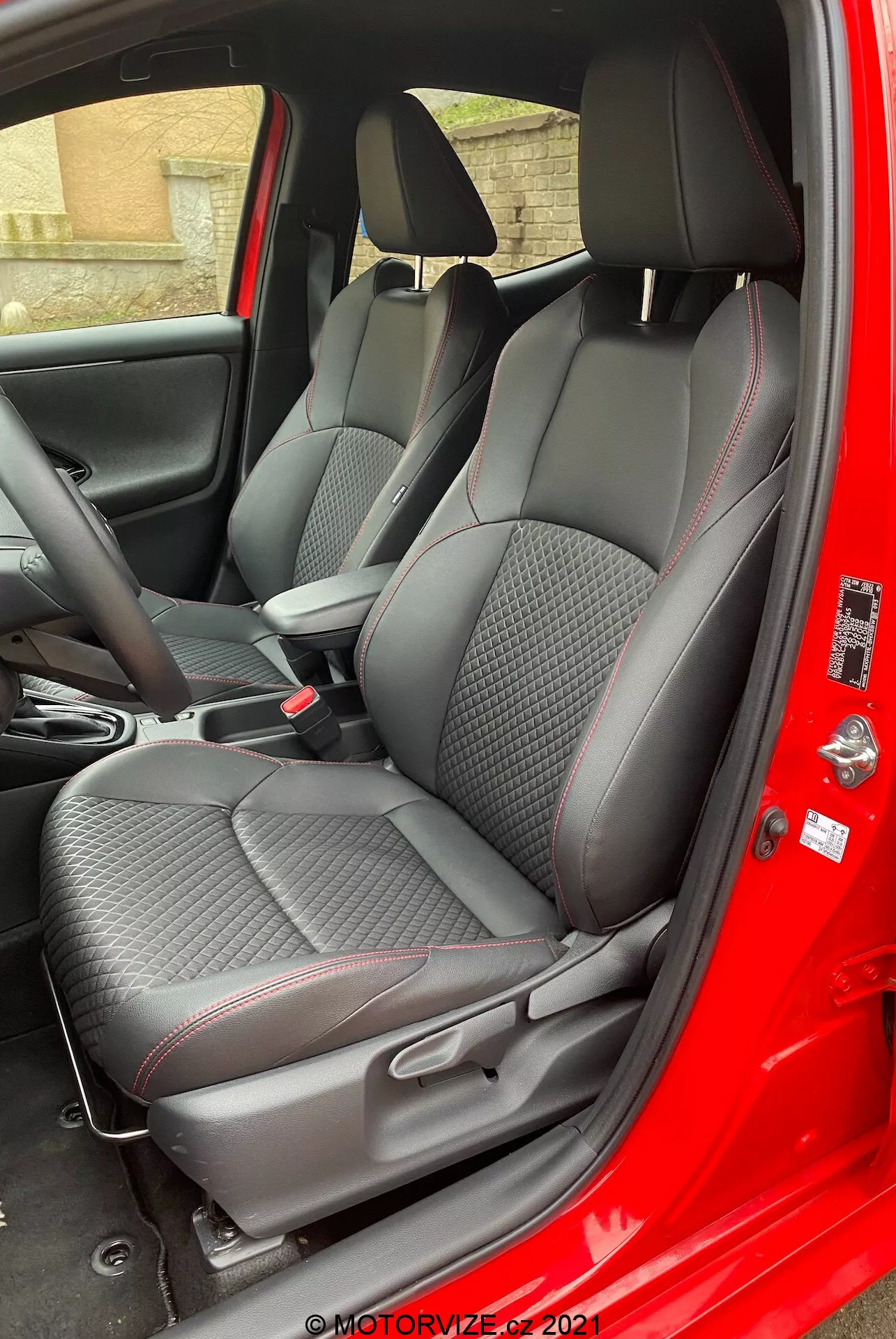Interiér modelu Toyota Yaris 2020 na pohled z místa spolujezdce s otevřenými dveřmi, zvýrazňující prémiové textilní sedačky se vzorem diamantu a červeným akcentováním, nastavitelné opěrky hlavy, ovládací panel středové konzoly a dveřní panel s texturou karbonových vláken a elektrickým ovládáním oken.