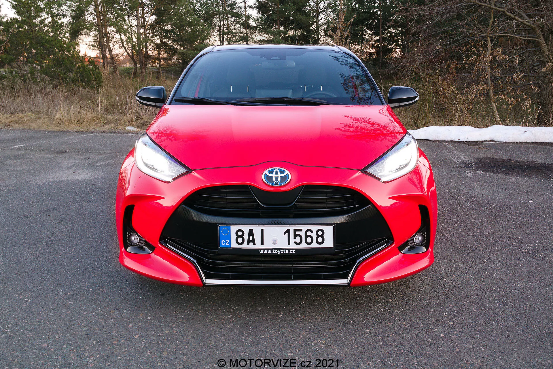 Obrázek ukazuje čelní pohled na červený Toyota Yaris 2020, pořízený pod mírným úhlem a zaměřující se na přední masku, světlomety a kapotu, s plně viditelnou registrační značkou. Vůz se vyznačuje charakteristickými designovými prvky, mezi které patří Toyota znak uprostřed mřížky, ostré hranaté světlomety zužující se ke mřížce a složité křivky na kapotě, které ukazují jeho aerodynamický a sportovní design. Fotografie pořízená během dne před soumrakem naznačuje, že vůz stojí na rovném povrchu v prostředí, které by mohlo být pozdní podzim nebo časné jaro.