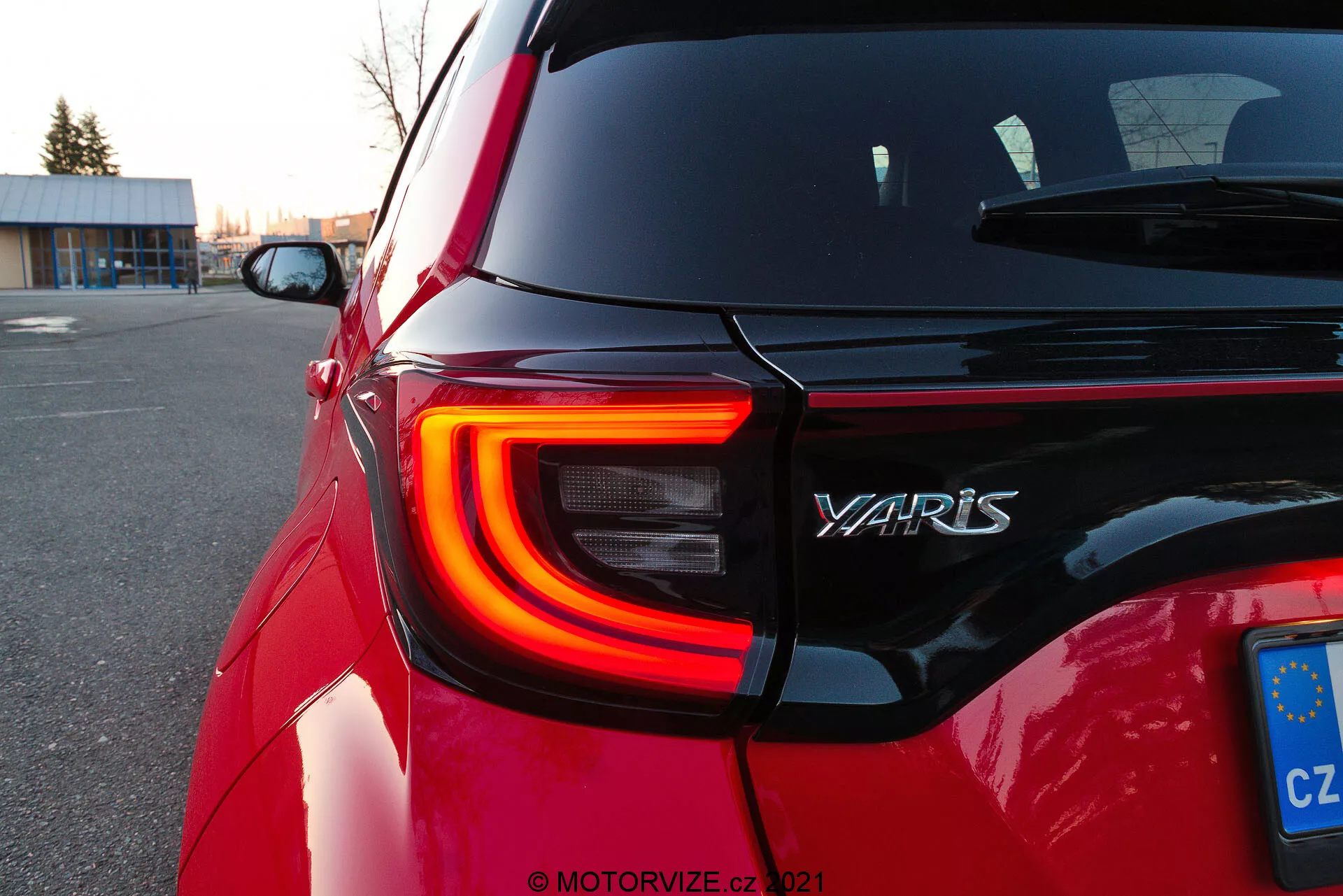 Detailní záběr na zadní část červeného modelu Toyota Yaris z roku 2020, zobrazující levé zadní LED světlo s výrazným tvarem C, název modelu 'YARIS' na zadních dveřích a poznávací značku Evropské unie z České republiky. Obraz v šikmém úhlu během soumraku zvýrazňuje obrysy a lesk auta, s rozmazaným pozadím, které soustředí pozornost na detaily designu.