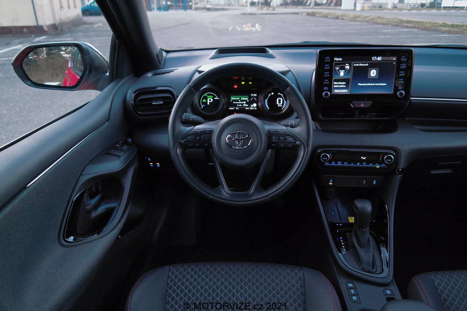 Interiérová perspektiva z řidičova sedadla Toyoty Yaris 2020, zobrazující volant s namontovanými ovládacími prvky, digitální přístrojový panel a infotainmentový displej. Interiér má ovládací tlačítka klimatu, řadicí páku a převážně černé barevné schéma s texturovanými povrchy a chromovými akcenty.