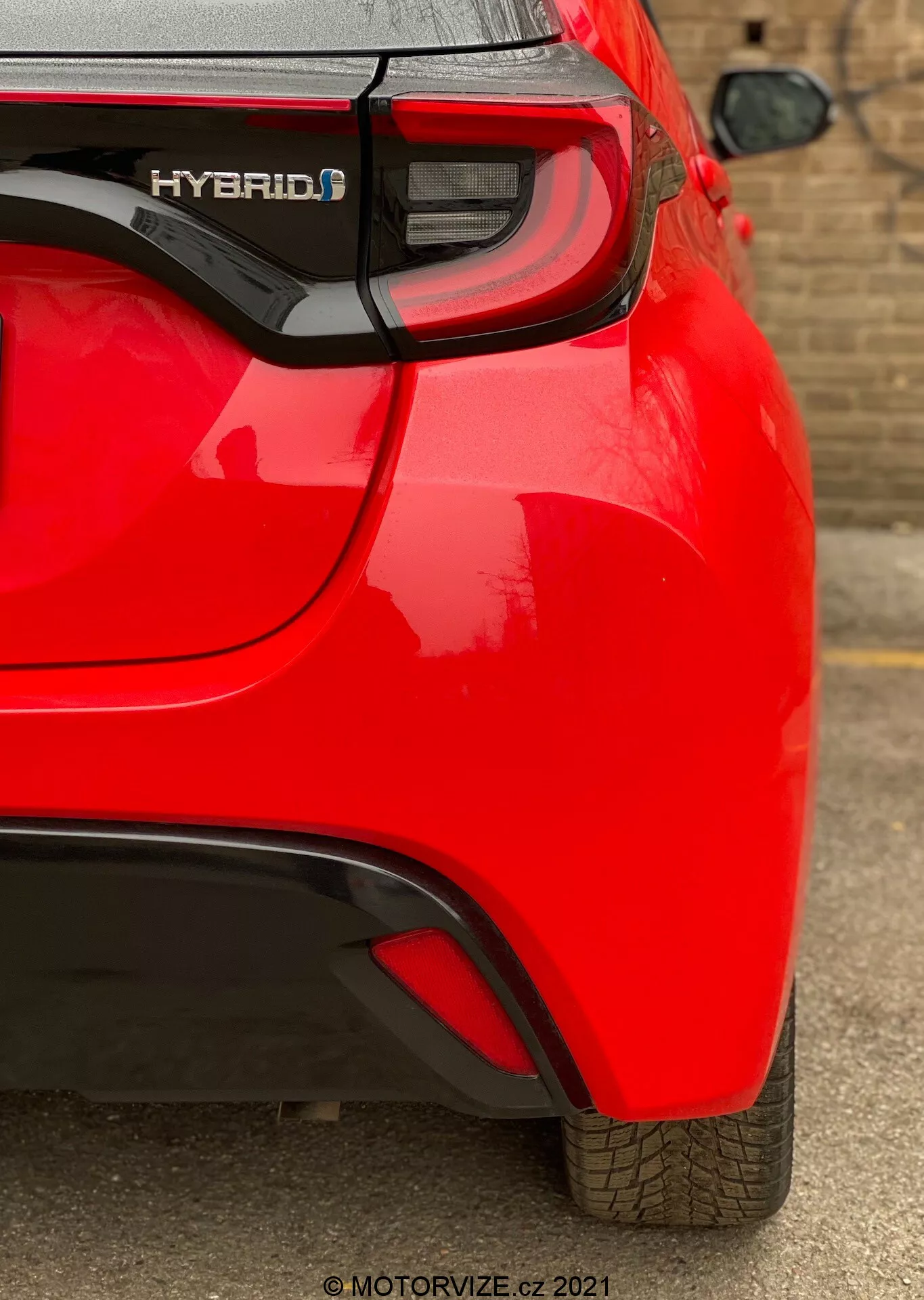 Detailní pohled na zadní část červeného hybridního vozu Toyota Yaris 2020 z úhlu, zaměřený na pravou zadní čtvrtinu. Zachycuje prvky jako označení 'HYBRID', moderní LED zadní světla, jemný aerodynamický záhyb na zadním nárazníku a viditelný parkovací senzor. Lesklý lak a dobře udržovaný vzorek pneumatiky naznačují, že vůz je nový nebo dobře udržovaný.