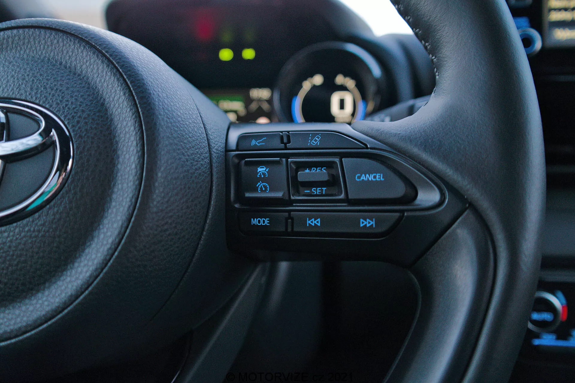 Záběr interiéru vozu Toyota Yaris 2020 z pohledu řidiče, zaměřený na levou stranu multifunkčního volantu. Hmatová tlačítka, včetně ikony bezpečnostního prvku, tempomatu s označením 'SET', tlačítka pro zrušení, 'MODE' pro multimédia a ovládání hlasitosti '+', '-', jsou jasně viditelné. Vozidlo je v režimu 'P' parkování, s přístrojovou deskou a pozadím mírně rozmazaným.