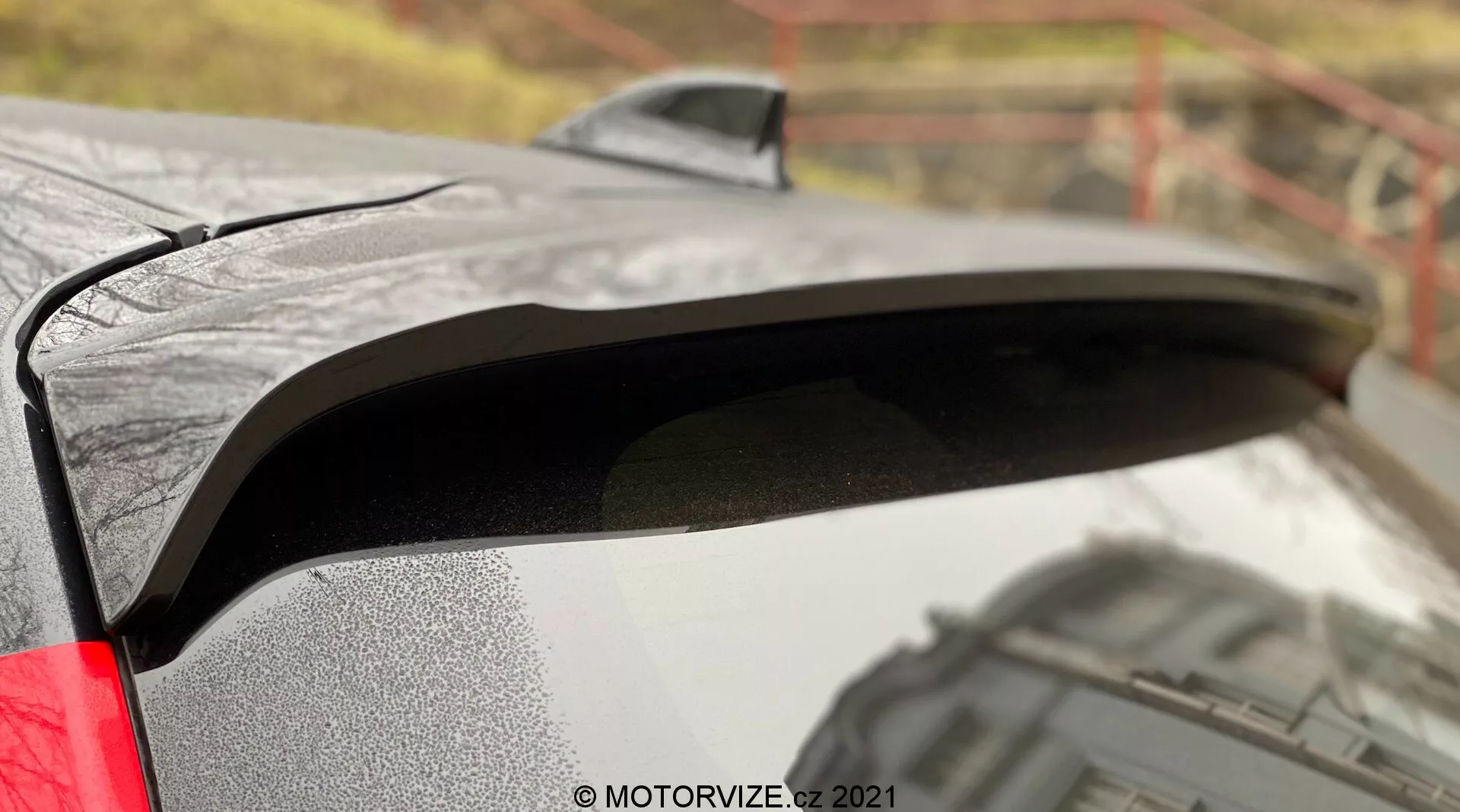 Detailní pohled na zadní spoiler Toyoty Yaris 2020 s černým lakem, který kontrastuje s metalickým povrchem karoserie. Obrázek zachycuje kapky vody na spoileru, což naznačuje mokré podmínky, a boční profil odhalující mírně nadzvednutý design pro zvýšenou aerodynamiku. Rozmazané venkovní pozadí dodává kontext k prémiovému laku vozu a viditelnému LED zadnímu světlu, což naznačuje moderní prvky.