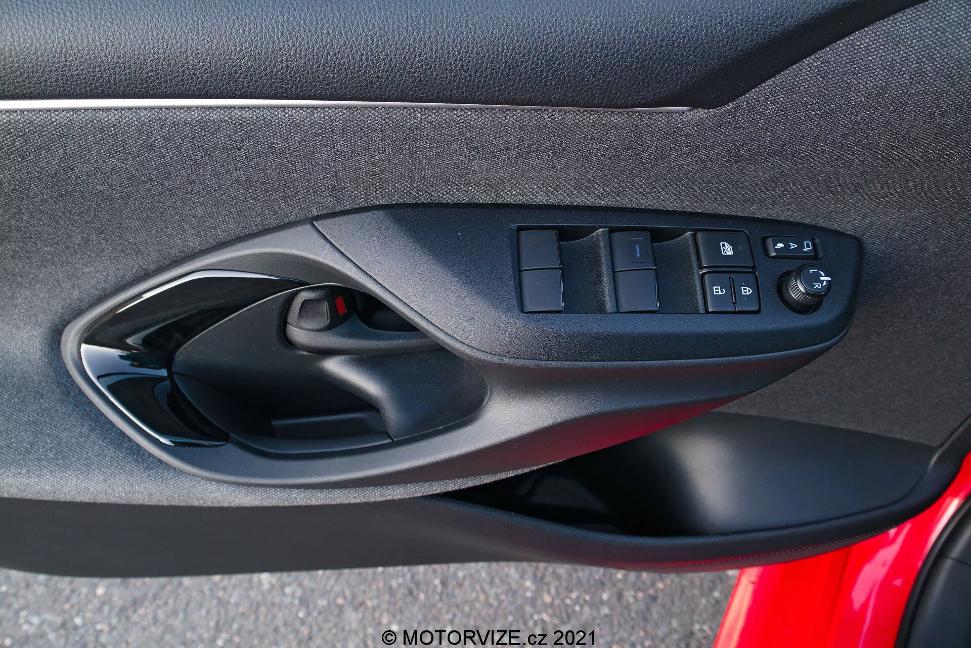 Interiérový dveřní panel Toyoty Yaris 2020, zaměřující se na ergonomicky navržené ovladače na straně řidiče, včetně vypínačů elektrických oken, tlačítka pro zamykání dveří a ovladačů zrcátek s elektrickým nastavením, pořízený z perspektivy sedícího cestujícího ve vozidle. Fotografie zdůrazňuje směs měkkých tkanin nebo kompozitních materiálů, černé barevné schéma a viditelnou mřížku reproduktoru jako součást audiosystému vozu.