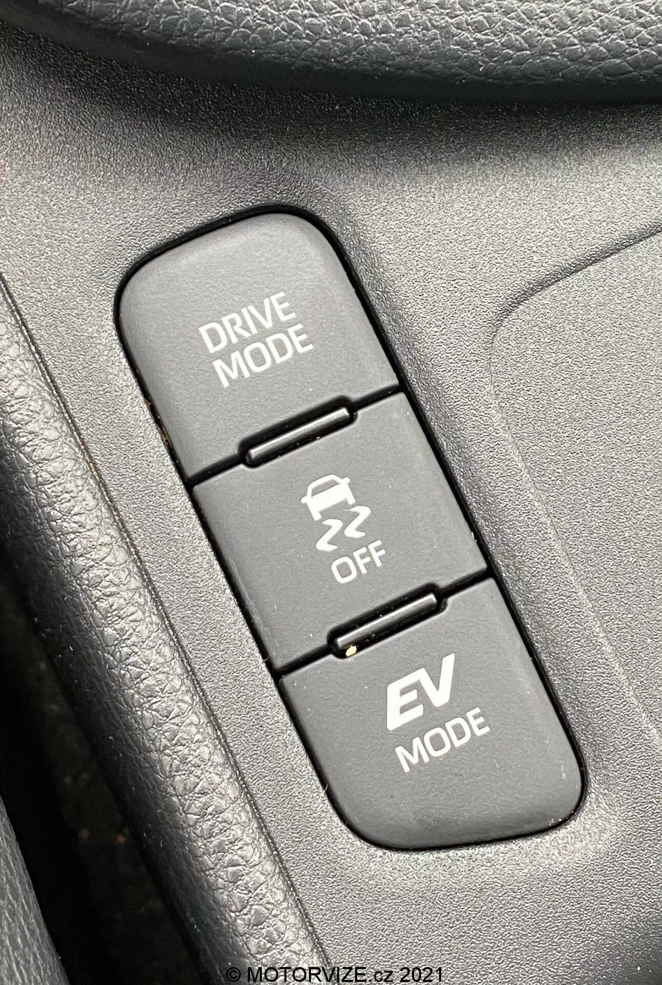 Detailní záběr interiéru vozu Toyota Yaris 2020, zaměřující se na tři vertikálně uspořádaná tlačítka ovládání jízdních režimů. Shora dolů: 'DRIVE MODE' s ikonou klikaté cesty, 'Traction Control OFF' s ikonou klouzajícího auta a 'EV MODE' označující funkci elektromobilu. Tlačítka jsou namontována na texturovaném povrchu, pravděpodobně části dveřního panelu nebo středové konzole. Obraz ostře zaostřuje na tlačítka, zatímco okolní textury jsou mírně rozostřené, a je osvětlen rovnoměrným denním světlem pro jasnou viditelnost.