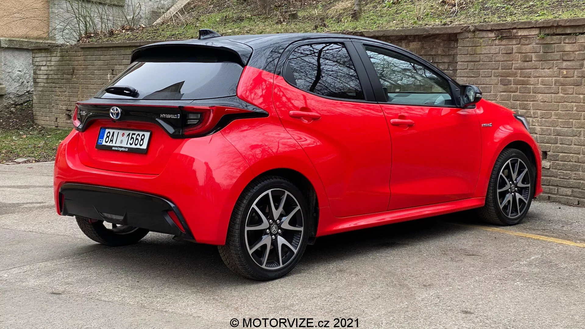 Tříčtvrteční zadní pohled na jasně červený, moderní hatchback s ostrými liniemi a záhyby, střešní spoilerem, LED zadními světly, žraločí anténou, vícerazradicovými koly a umístěný na parkovišti s reflexním, dobře udržovaným nátěrem, což naznačuje nedávný nebo dobře udržovaný model v sportovní úpravě.