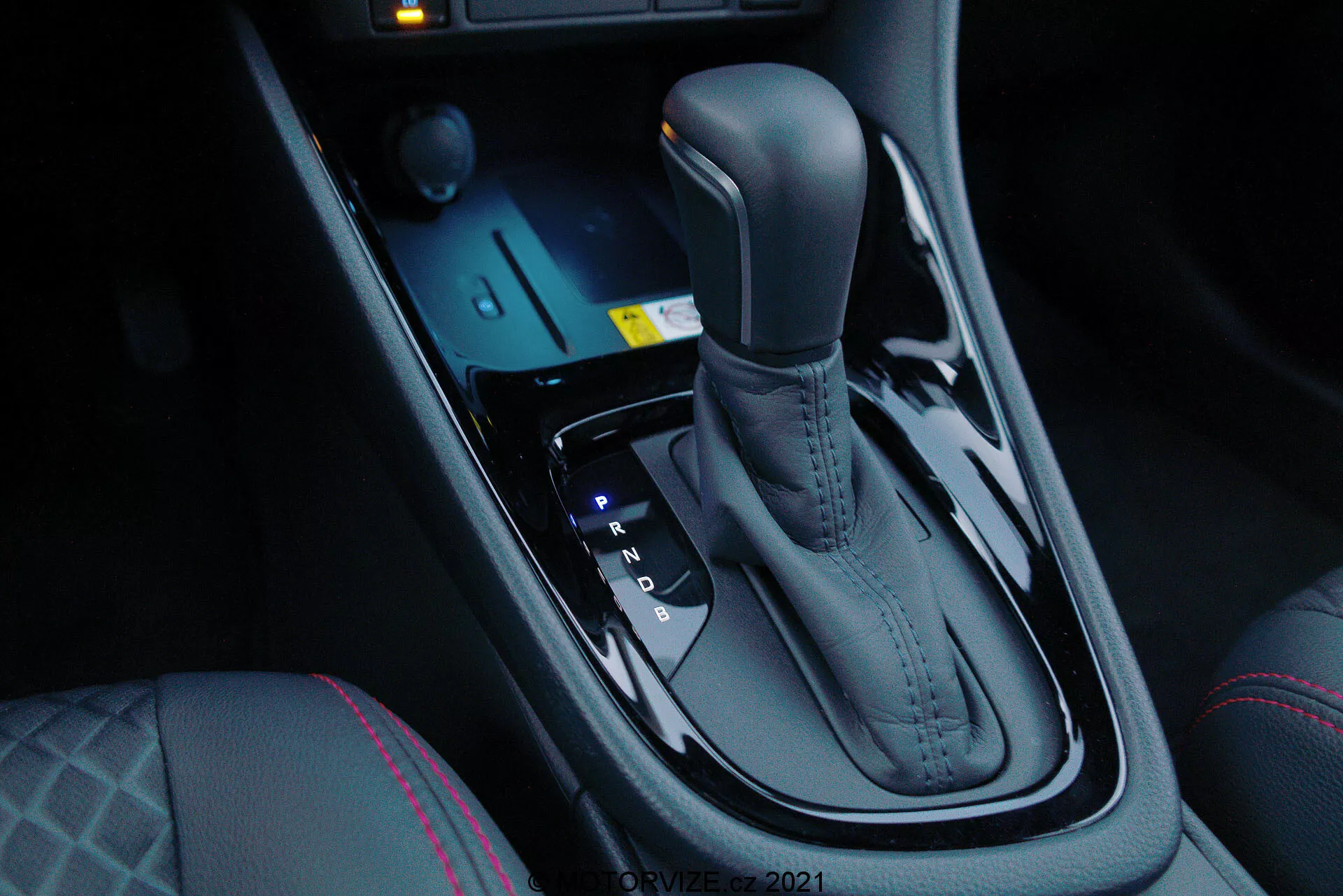 Detailní pohled na automatickou převodovku modelu Toyota Yaris 2020, viděný ze strany spolujezdce ve sklonu dolů, který zdůrazňuje elegantní kovový povrch a ergonomický design. Převodovka je umístěna uprostřed mezi sedadly řidiče a spolujezdce a zahrnuje polohy Park, Reverse, Neutral a Drive, stejně jako možnosti pro ruční řazení (+ a -). Před převodovkou jsou elektronické ovladače pro jízdní režimy a kontrolu trakce společně s modře osvětleným startovacím tlačítkem. Tmavá, lesklá středová konzola přidává moderní luxus, který doplňují ovladače vyhřívání sedadel a výrazné švy na řidičově sedadle, což indikuje zvýšený komfort a pokročilé jízdní dynamiky.
