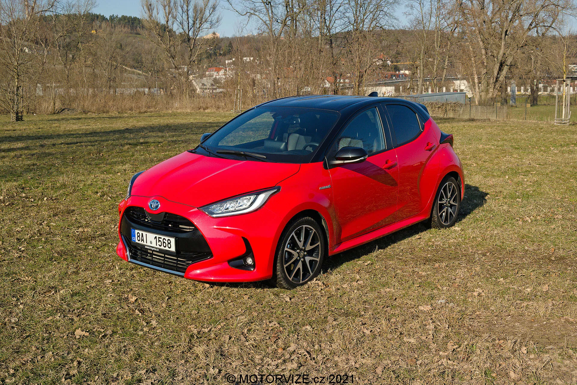 Obrázek zobrazuje jasně červený Toyota Yaris 2020, zaparkovaný na nerovném travnatém terénu v polovenkovské oblasti, pohled z tříčtvrteční perspektivy. Moderní, aerodynamický design vozu zahrnuje ostré úhly kolem světlometů a kompaktní, sportovní vzhled. Viditelné jsou tmavá vícepaprsková kola a stylizované LED světlomety. Boční okna a přední čelní sklo poskytují tmavý náhled do interiéru, což naznačuje možnost pokročilých automobilových systémů.