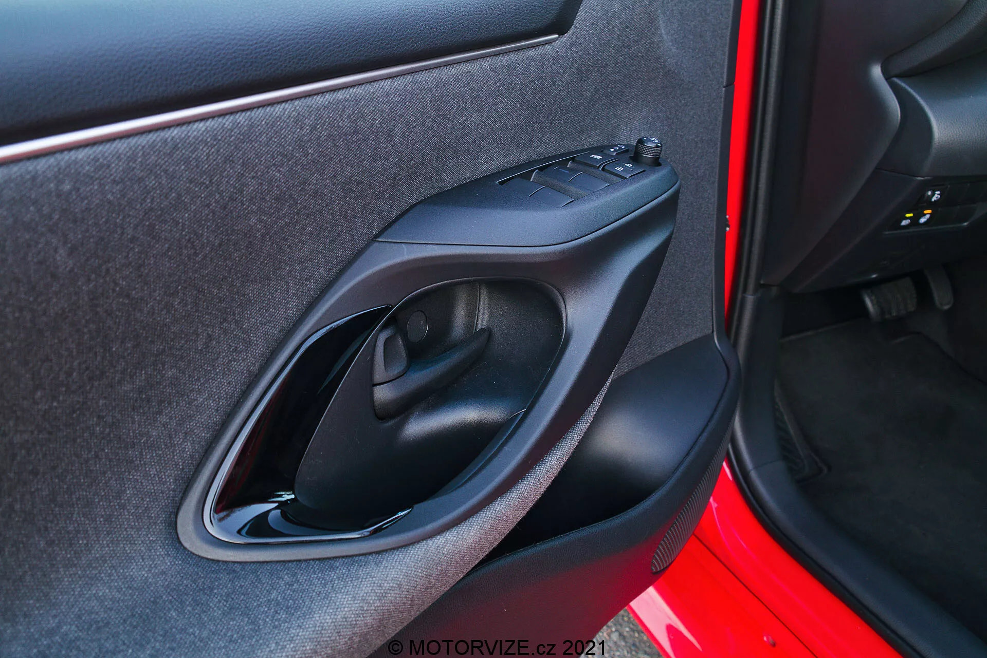 Zblízka záběr interiérového panelu dveří vozu Toyota Yaris 2020 pořízený z exteriéru. Snímek se zaměřuje na panel dveří na pravé straně, který obsahuje designové a technologické prvky, jako je matně černé plastové madlo dveří, ovládací panel pro elektřinou ovládaná okna, ergonomicky tvarovanou opěrku pro ruce a malou úložnou kapsu. Exteriér dveří je lakován sytou červenou barvou.