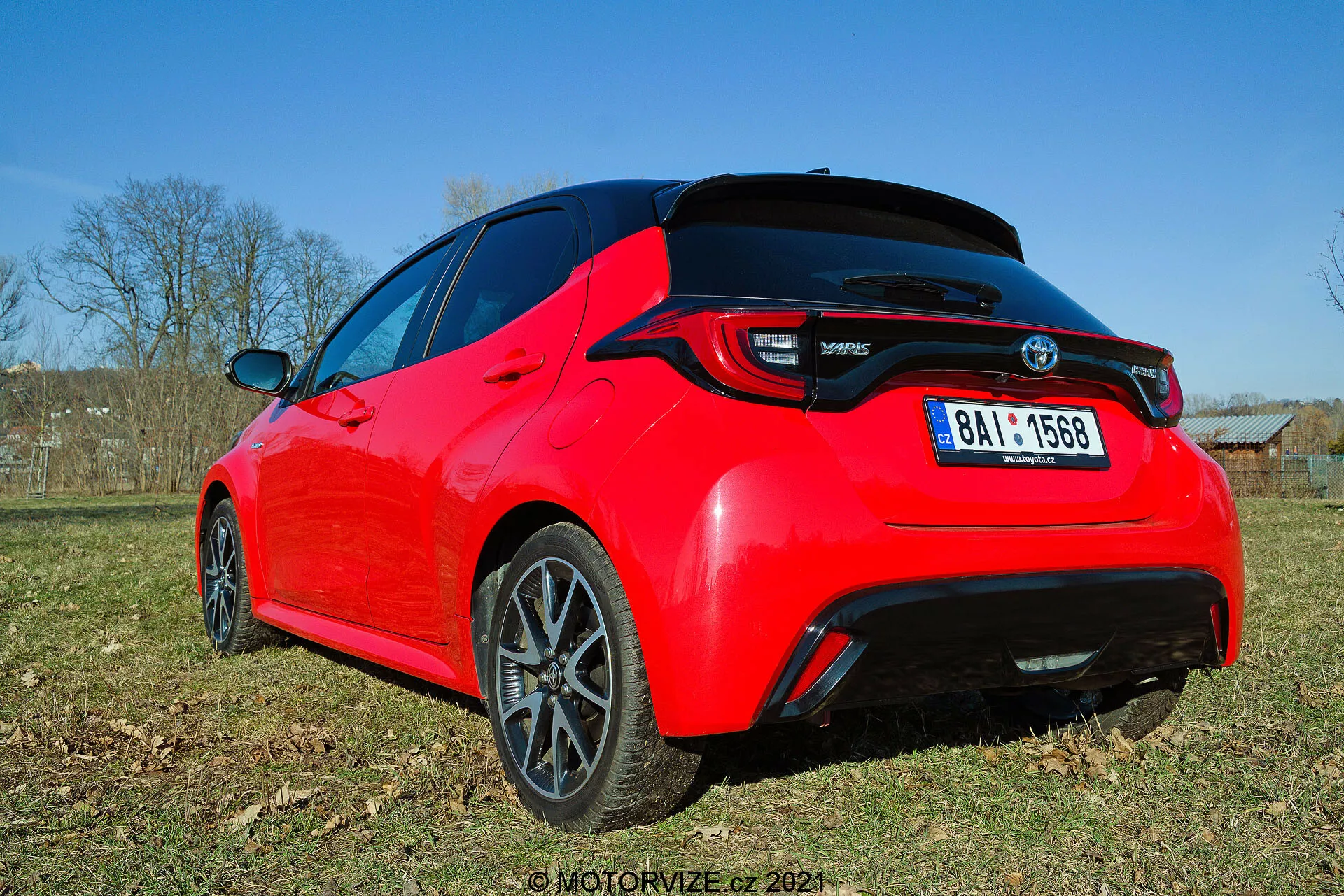 Zadní tři čtvrtiny pohled na jasně červený hatchback Toyota Yaris 2020, viděn z nízkého úhlu, zdůrazňující designové linie a klíčové prvky, jako jsou LED zadní světla, zadní spoiler, anténa ve tvaru žraločí ploutve a tmavě dokončená multi-spoke slitá kola. Fotografie zachycuje detail zadní části a částečně bočního profilu za denního světla, což zvýrazňuje jeho sportovní estetiku a moderní prvky.