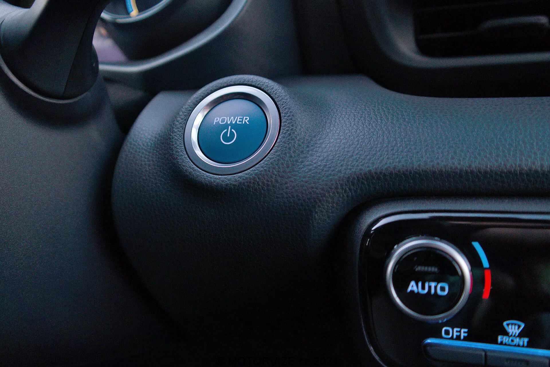 Pohled na interiér ze strany řidiče Toyota Yaris 2020, který zvýrazňuje tlačítko start/stop a ovládání klimatizace. Tlačítko Power s modrým osvětleným symbolem je výrazné v levém horním rohu se stříbrným akcentem. Níže a napravo se nachází ovládací knoflíky klimatizace, které ukazují modré světlo a symbol vločky pro chladnou teplotu, s označeními 'AUTO', 'OFF' a 'FRONT'. Smíšené textury a hladké černé syntetické materiály naznačují minimalistický styl s funkcemi bezklíčového startu a automatického ovládání klimatizace.