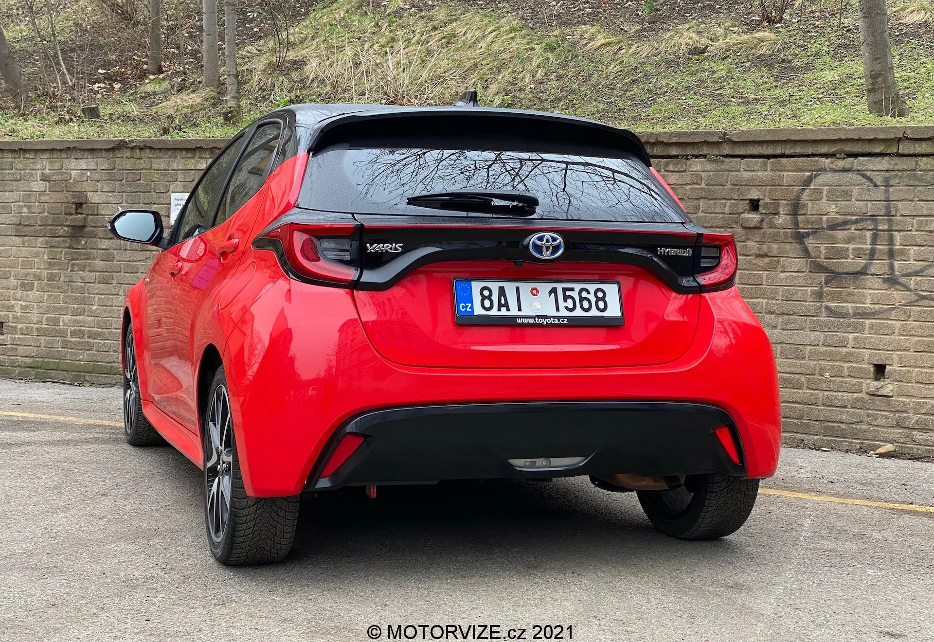 Vysoce kvalitní snímek červeného Toyota Yaris 2020 hatchbacku zobrazeného ze zadního tříčtvrtečního úhlu. Vozidlo se vyznačuje výraznými, šikmými zadními světly, centrálně umístěným emblémem Toyota nad pravděpodobnou zpětnou kamerou a modelem 'Yaris' označeným na spodní části levých dveří kufru. Pod oblastí registrační značky je černý ozdobný prvek, který by mohl být z plastu nebo uhlíkových vláken, a malý zadní spoiler se táhne nad zadním oknem. Auto zaparkované poblíž obrubníku na dlážděné silnici vedle cihlové zdi naznačuje městské nebo rezidenční prostředí.