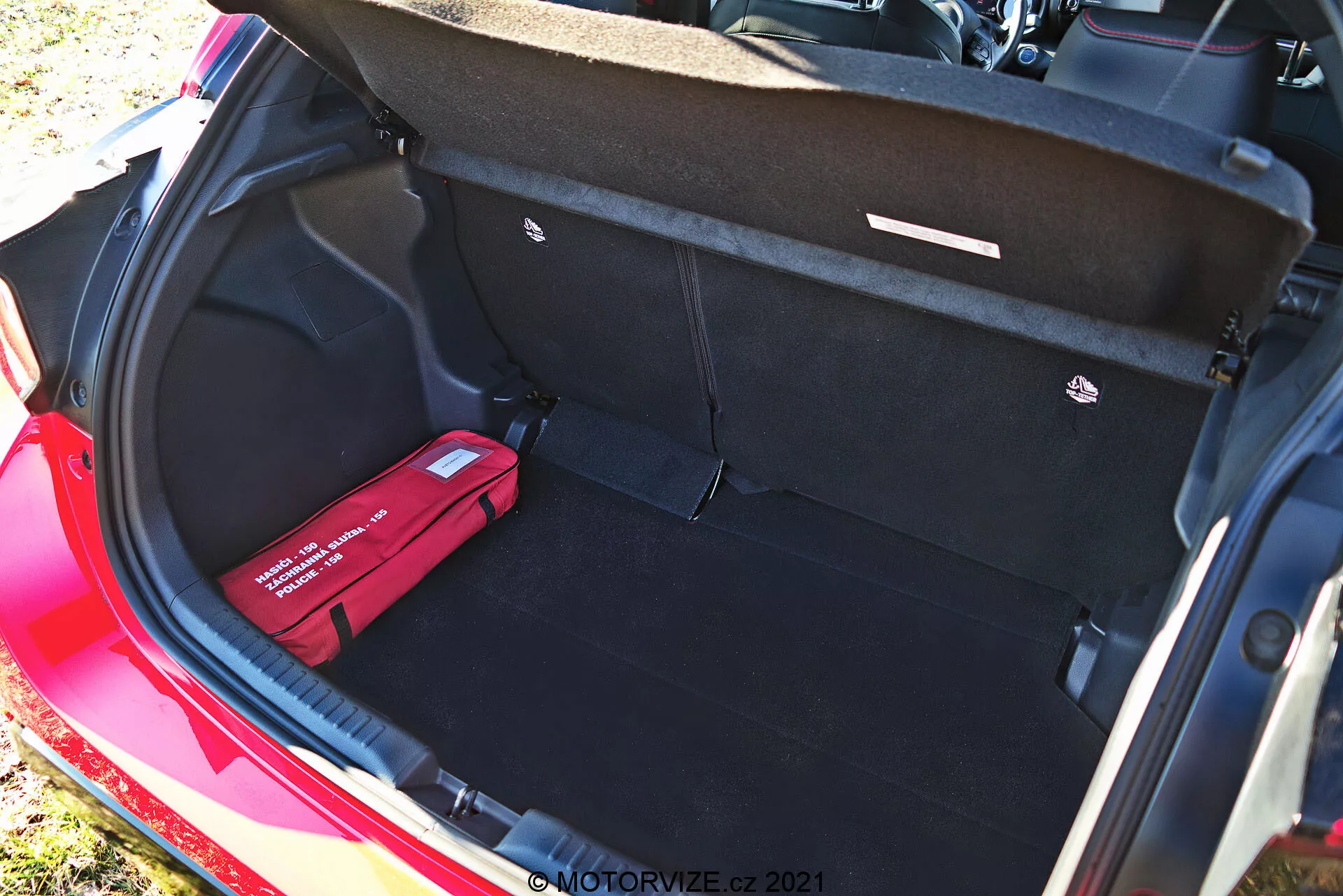 Zavazadlový prostor červeného Toyota Yaris 2020 pohledem z vyššího úhlu, který ukazuje jeho čistý, prostorný interiér s černým kobercem, otevřeným víkem kufru s táhly, plochou nákladovou podlahou, háčkem na náklad vpravo a červenou lékárničkou nebo nouzovou sadou vlevo. Tento denní snímek zdůrazňuje dostatečný úložný prostor a organizační prvky vozidla.