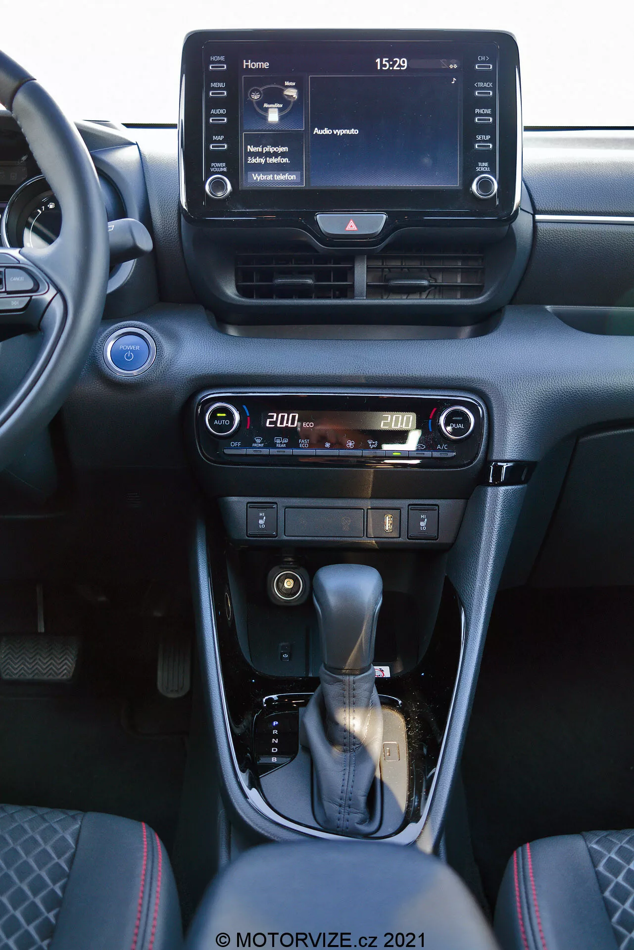 Pohled na interiér Toyota Yaris 2020 zaměřený na středovou konzoli a prvky přístrojové desky. Fotografie pořízená ze sedadla předního spolujezdce zobrazuje digitální infotainment systém s domovskou obrazovkou s časem 15:29 a různými menu možnostmi, centrální ventilace a snadno použitelné ovladače klimatizace a topení s digitálním displejem teploty nastavené na 20,0 stupňů. Středová konzole obsahuje koženou řadicí páku se stříbrným akcentem a ruční parkovací brzdu. Interiér je navržen v tmavých tónech s kovovými doplňky, což poskytuje moderní a sofistikovaný vzhled.