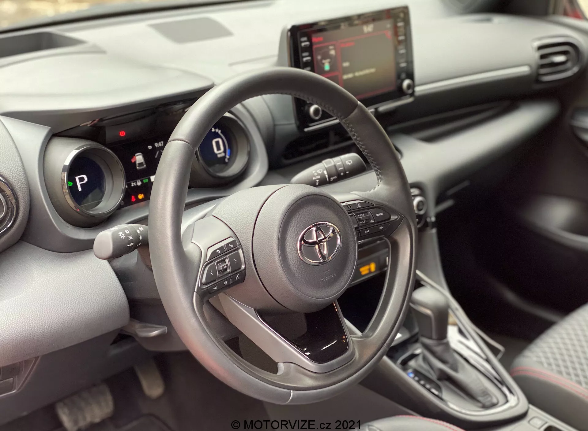 Pohled na interiér Toyoty Yaris 2020 z místa spolujezdce, zaměřený na řidičovu oblast. Obrázek zvýrazňuje tříramenný volant Toyota s ovládacími tlačítky pro audio, Bluetooth a tempomat, s emblémem Toyota uprostřed. Za volantem je částečně viditelný digitální přístrojový panel, který ukazuje, že auto je v režimu 'P' (parkovací). Středová konzola zobrazuje infotainment obrazovku a pod ní ovládání HVAC, spolu s možnými USB nebo aux vstupy. Obrázek také ukazuje automatickou převodovku s moderním designem a krytem z materiálu podobného kůži. Interiér má moderní design s kombinací měkkých materiálů a stříbrných akcentů, spolu s držáky nápojů nebo malými úložnými přihrádkami vedle řadicí páky. Částečně viditelný panel dveří odhaluje ovládací panel oken, což naznačuje elektrická okna.