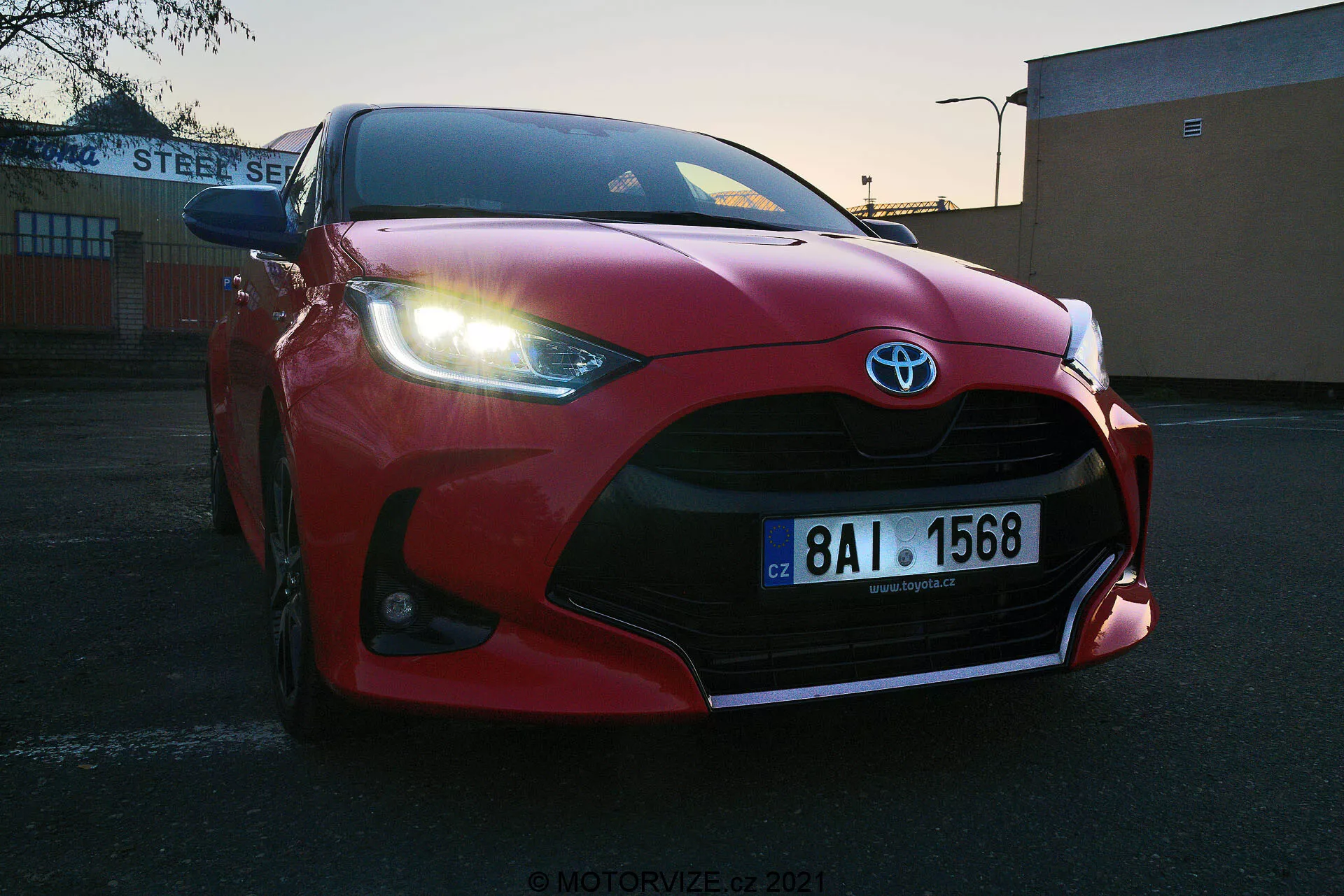 Nízký úhel zblízka červeného vozu Toyota Yaris 2020 zaparkovaného venku při východu nebo západu slunce. Zdůrazněná přední maska, nárazník a ostře tvarované LED světlomety s výraznými přívody vzduchu zdůrazňujícími znak Toyota. Viditelná evropská registrační značka, umístěná na pozadí průmyslové nebo komerční budovy s nápisem 'STEEL SERVICE'. Jasná obloha s přechodovým osvětlením zvýrazňuje sportovní design vozidla.