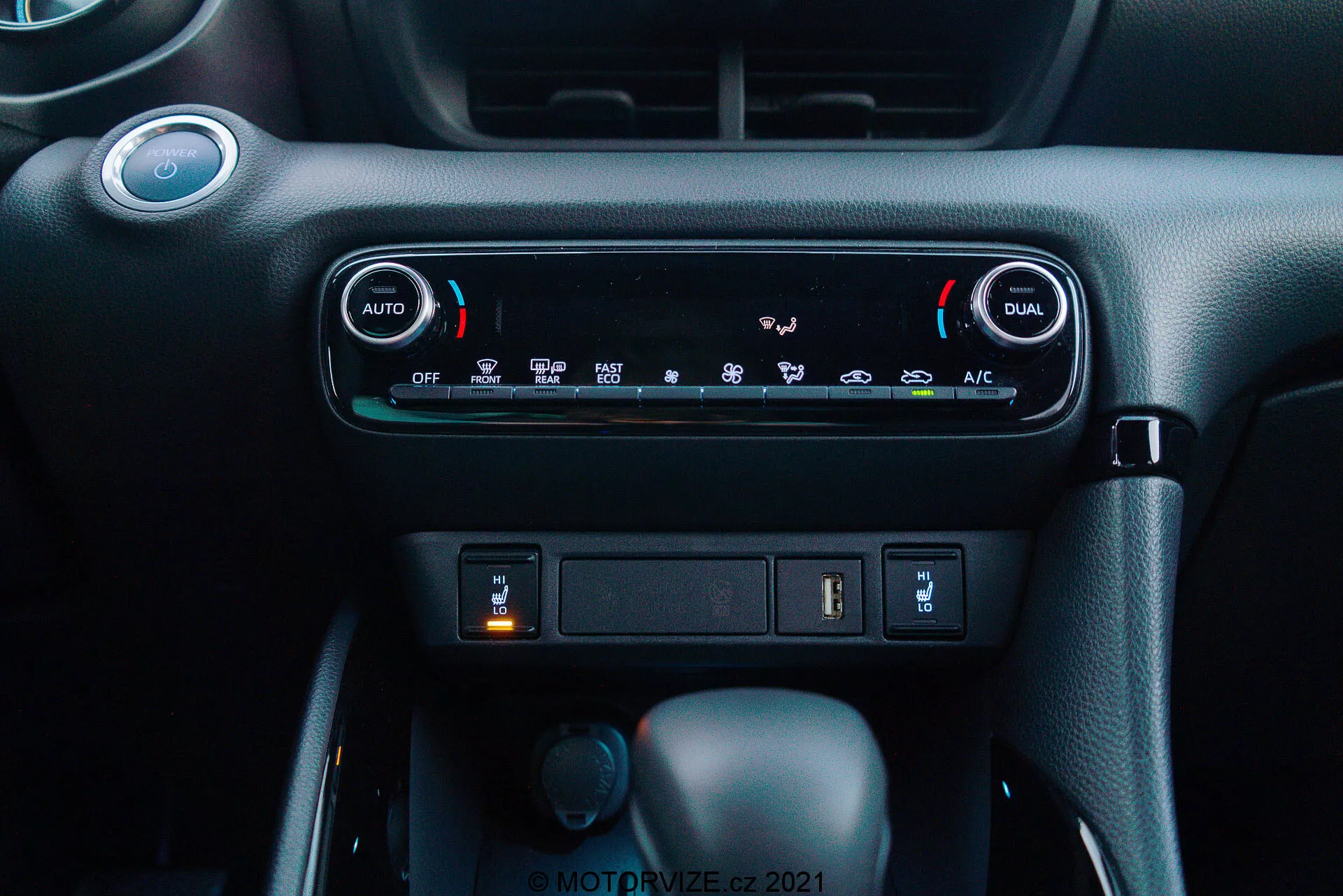 Detailní pohled na přístrojovou desku Toyota Yaris 2020: Obrázek zvýrazňuje klimatizační systém s duální zónovou automatickou klimatizací, se zobrazenými tlačítky 'AUTO' a 'DUAL' pro nezávislé nastavení teploty pro řidiče a spolujezdce. Pod klimatizací je úložný slot s USB portem a potenciální 12V napájecí zásuvkou. Níže jsou vidět tři ovladače: centrální je řadicí páka v poloze 'Park', což naznačuje automatickou převodovku, menší otočný ovladač vlevo pravděpodobně pro výběr režimu jízdy a tlačítko vpravo možná pro elektronickou parkovací brzdu. Přístrojová deska má tmavé barevné schéma s prémiovým texturovaným povrchem, vytvářející moderní, uživatelsky přívětivé prostředí kabiny.