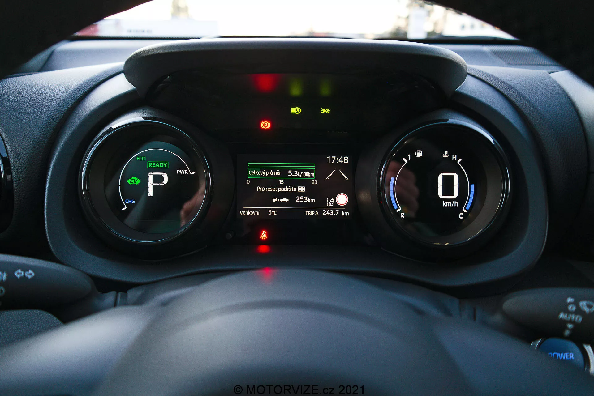 Pohled do interiéru Toyota Yaris 2020 zaměřený na přístrojový panel, pořízený z pohledu řidiče, zobrazující digitální přístrojovou desku, indikátory elektrického nebo hybridního pohonu a multifunkční displej se zobrazením jako je vzdálenost cesty, vnější teplota a zbývající dojezd baterie.