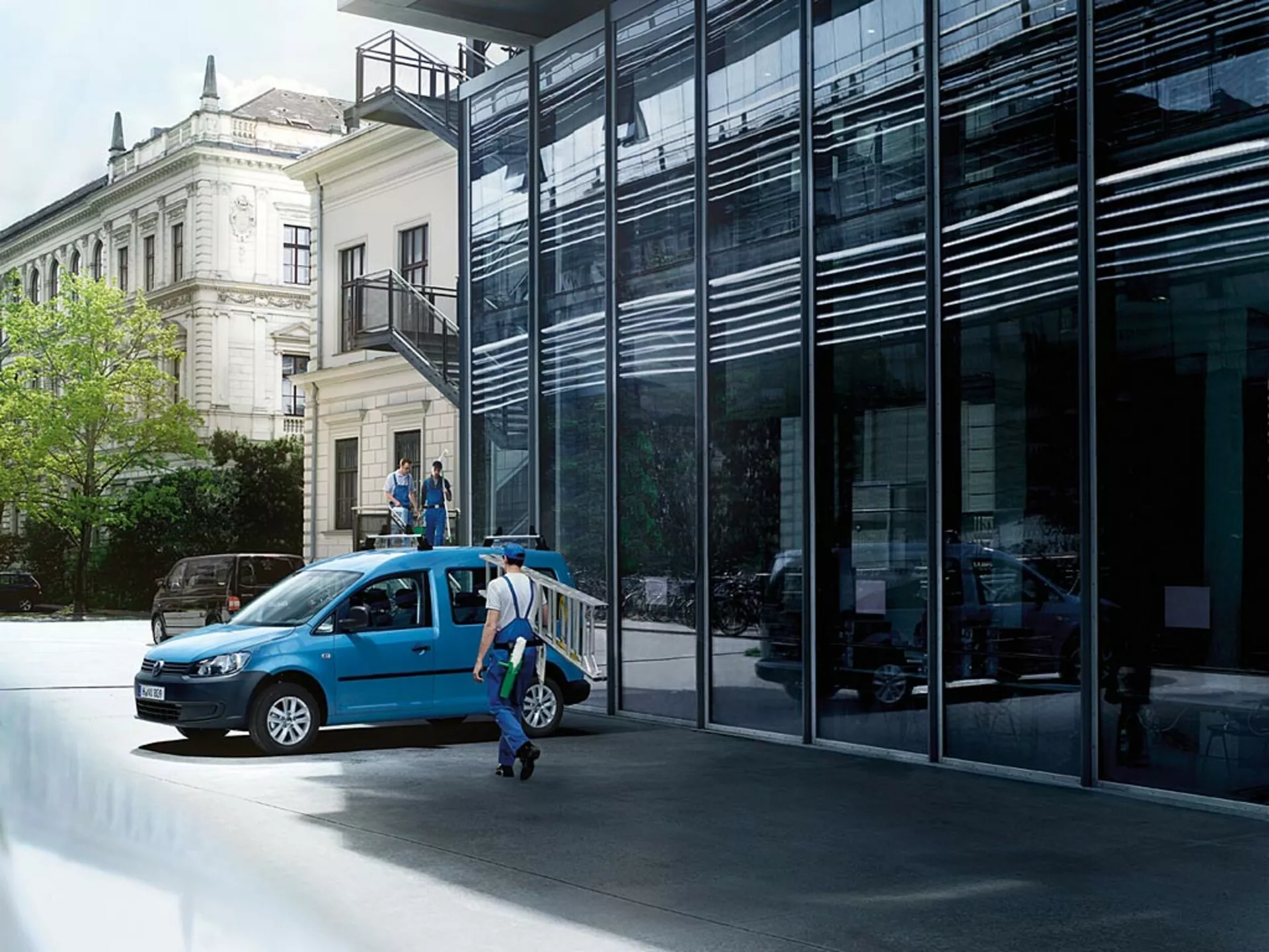Modrý VW Caddy rok 2010 parkovaný na městské ulici, zobrazený z čelně-bočního pohledu s otevřenými dveřmi a zdůrazněním přední mřížky, světlometů, kol a náhledu do interiéru na pozadí moderní a klasické architektury