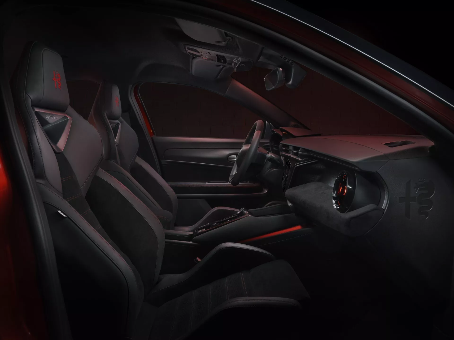 Interiérový pohled na Alfa Romeo Junior 2024 ukazující stranu řidiče s moderními, sportovně laděnými prvky včetně volantu s plochým dnem a integrovanými ovladači, elegantní středovou konzolí bez tradiční řadicí páky, sportovních sedadel s výraznými postranními opěrkami a luxusních materiálů s akcenty ambientního červeného osvětlení