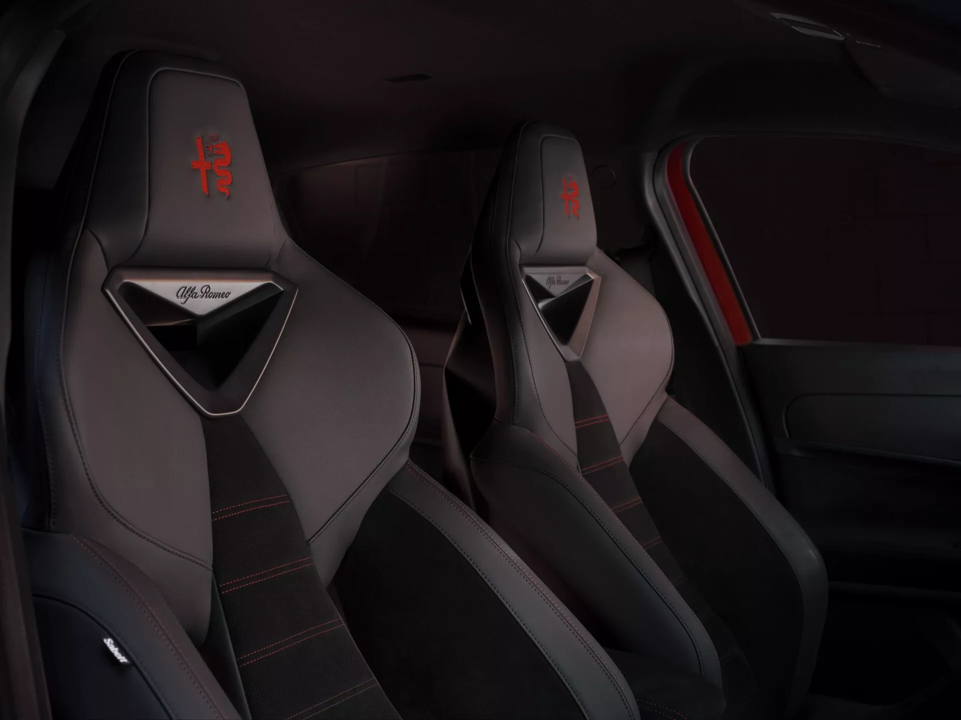 Pohled na interiér Alfa-Romeo Junior 2024 se zaměřením na sportovní přední sedadla s červeným prošíváním a logem, předvádějící kvalitní čalounění a sportovní designové prvky
