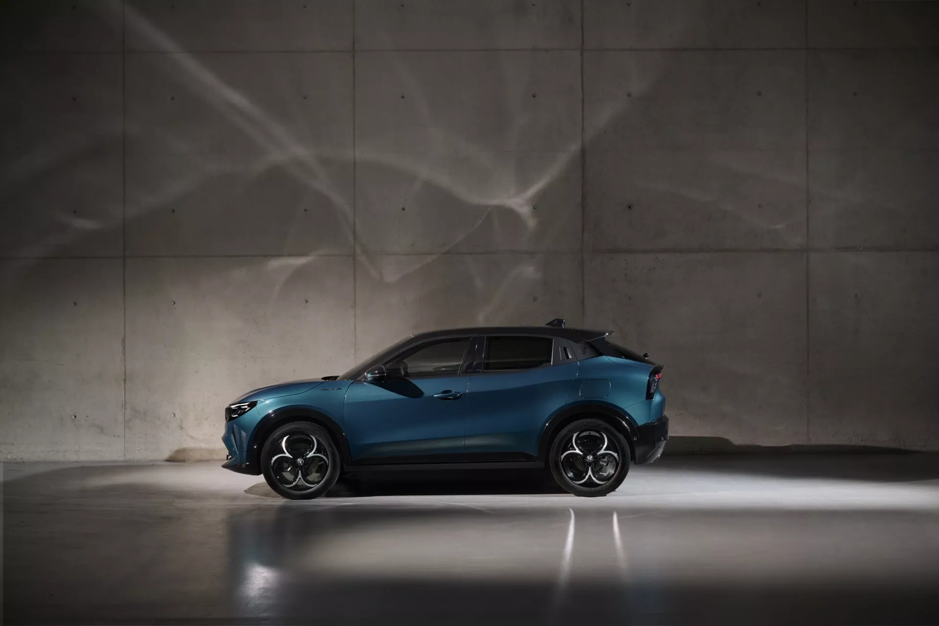 Alfa Romeo Junior 2024 v metalické modré barvě, pohled z profilu zdůrazňující sportovní a kompaktní design crossoveru, s velkými slitinovými koly a svažující se střechou, dramatičtím osvětlením a uměleckými stínovými vzory na pozadí