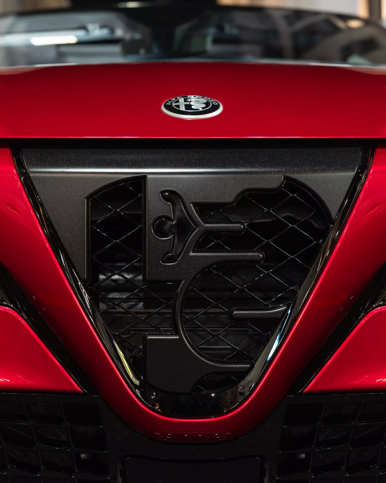 Detailní pohled na mřížku a logo výrobce Alfa-Romeo Junior (966) pro rok 2024, zdůrazňující složitý hexagonální síťový design, aerodynamické prvky a lesklý červený povrch